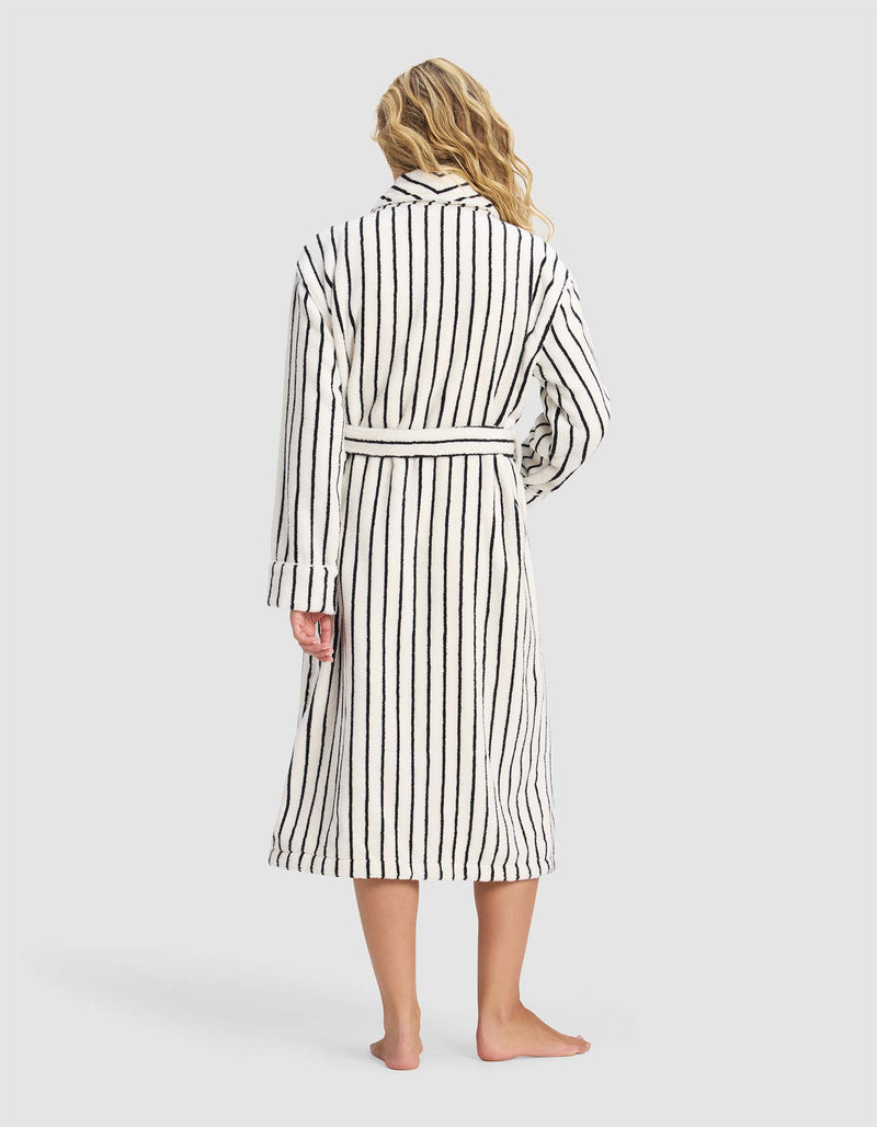 A person with long, wavy blonde hair stands barefoot facing away, wearing the Cozy Earth Striped Luxe Bath Robe against a plain light gray background.