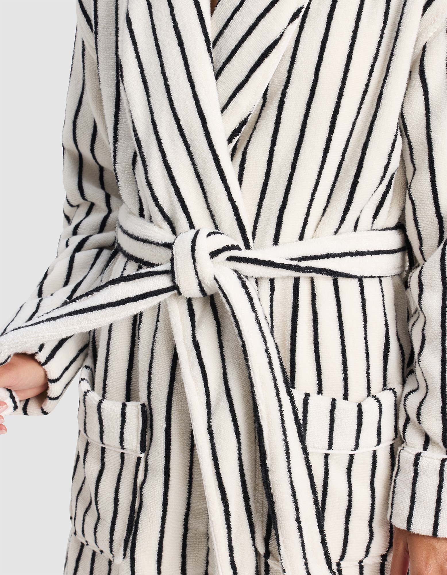 A person wears the Cozy Earth Luxe Bath Robe—plush, white with black stripes, tied at the waist with a matching belt. The robe features front pockets and the person holds one end of the belt in hand.