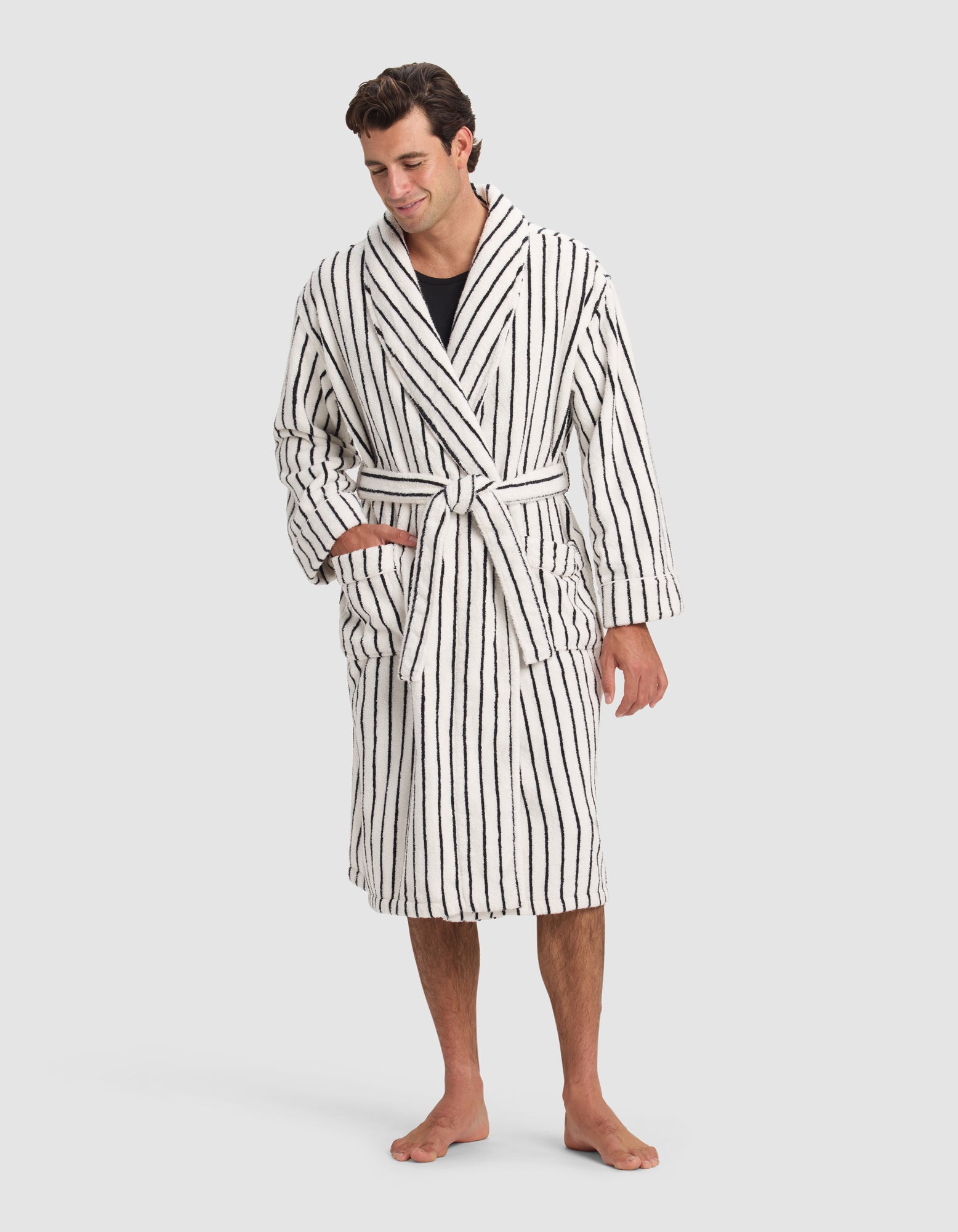 A man stands barefoot in the Luxe Bath Robe by Cozy Earth, featuring vertical black stripes, a matching belt, and pockets. He has short dark hair, smiles slightly, with one hand in his pocket and the other at his side.