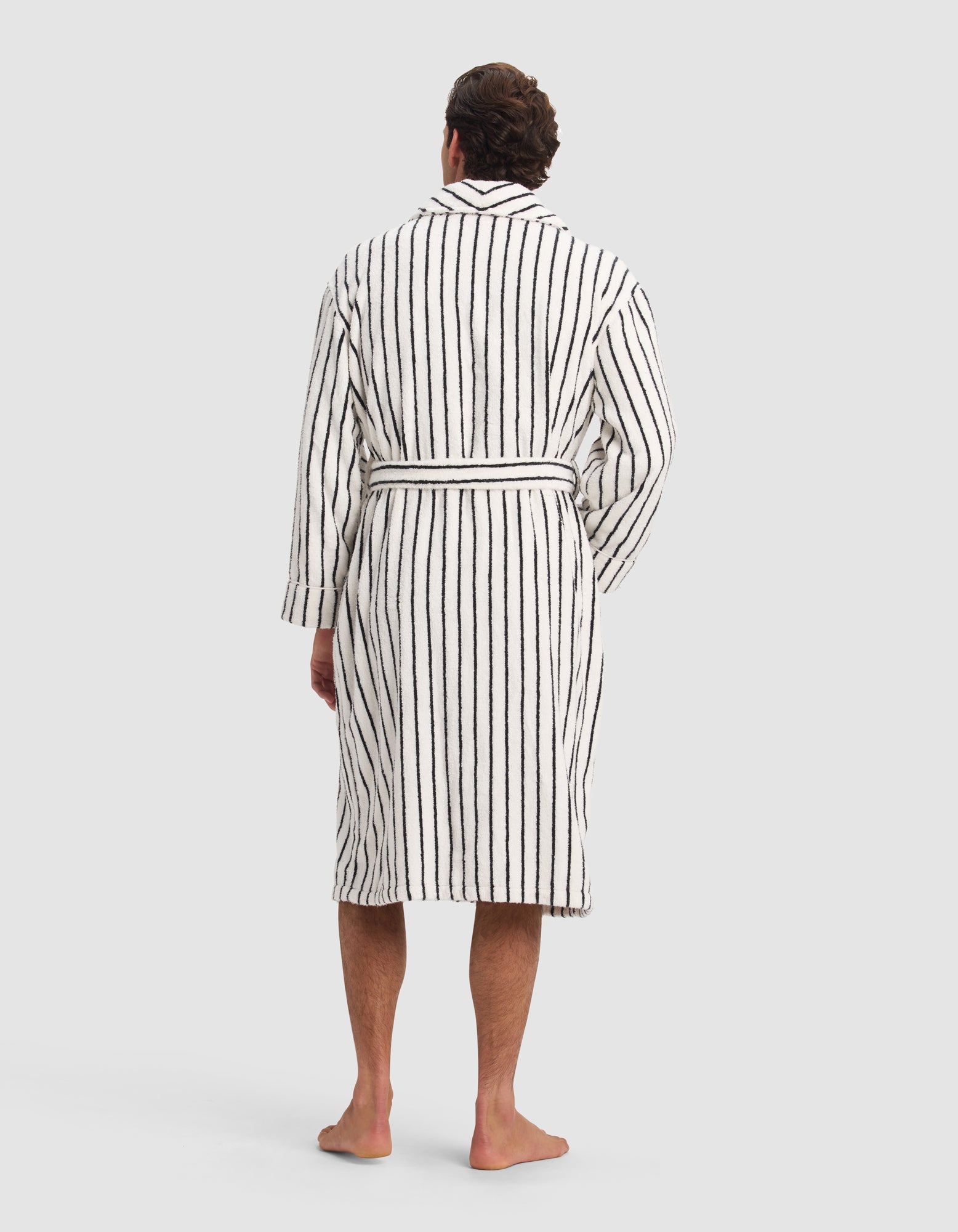 A person stands barefoot facing away, wearing the Luxe Bath Robe by Cozy Earth—a knee-length, white robe with vertical black stripes and a belt—against a plain light gray background.