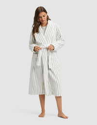 A woman, barefoot, wears the Cozy Earth Striped Luxe Bath Robe in long white fabric with vertical blue stripes, tying the waist belt as she looks downward with her long hair over one shoulder. |Color:Brook Stripe