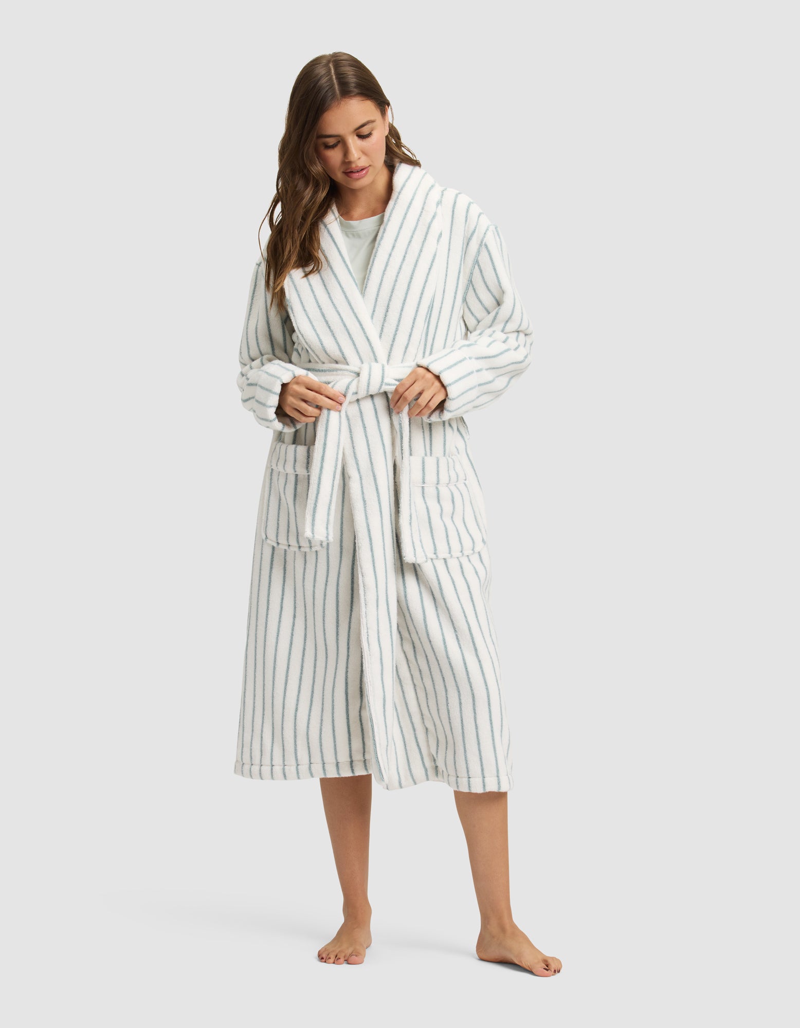 A woman, barefoot, wears the Cozy Earth Striped Luxe Bath Robe in long white fabric with vertical blue stripes, tying the waist belt as she looks downward with her long hair over one shoulder. 