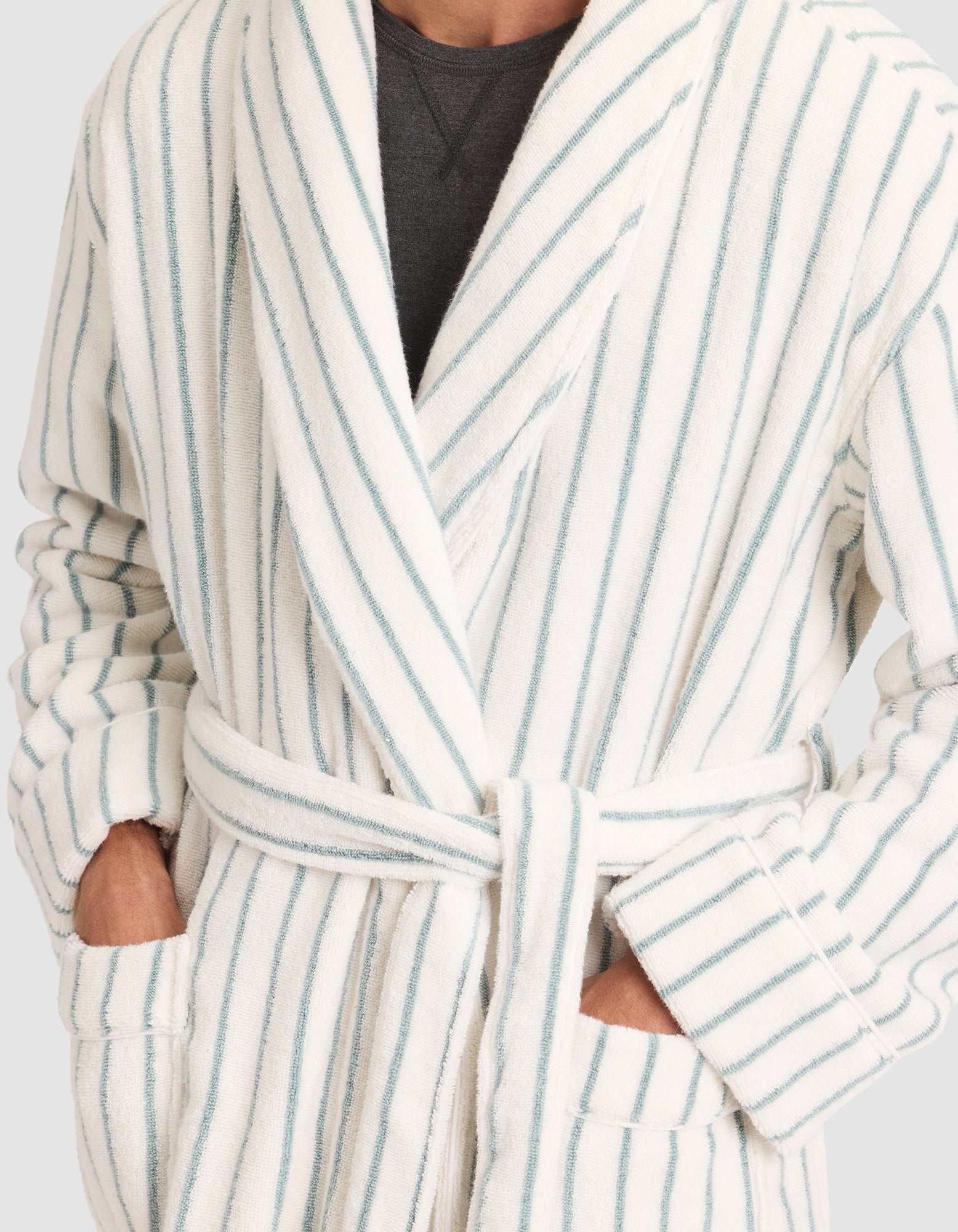 A person wearing the Cozy Earth Striped Luxe Bath Robe with light blue vertical stripes, belted at the waist, hands in pockets. Only the torso is shown, and a dark shirt is visible underneath. 