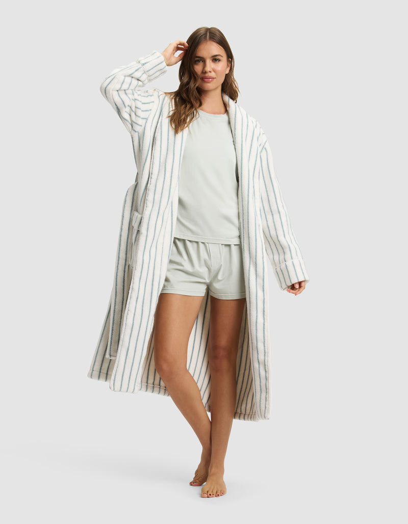 A woman stands barefoot, smiling with one hand in her hair, wearing the Cozy Earth Striped Luxe Bath Robe—a long, white robe with thin blue stripes—over a light-colored pajama set with shorts.