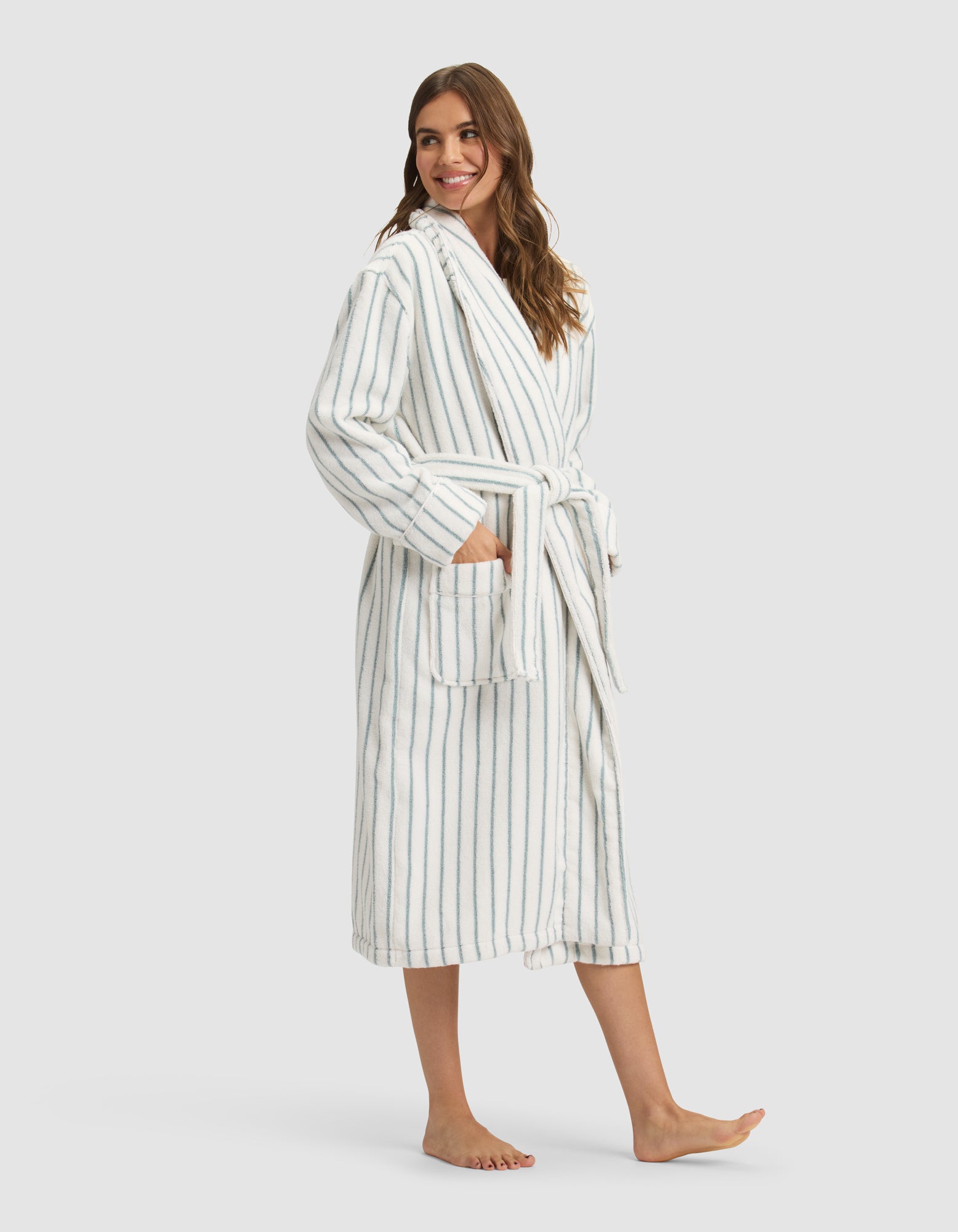 A woman stands barefoot, smiling, wearing the Cozy Earth Striped Luxe Bath Robe—long, white with blue vertical stripes, a matching belt at the waist, and large front pockets where she rests her hands. 