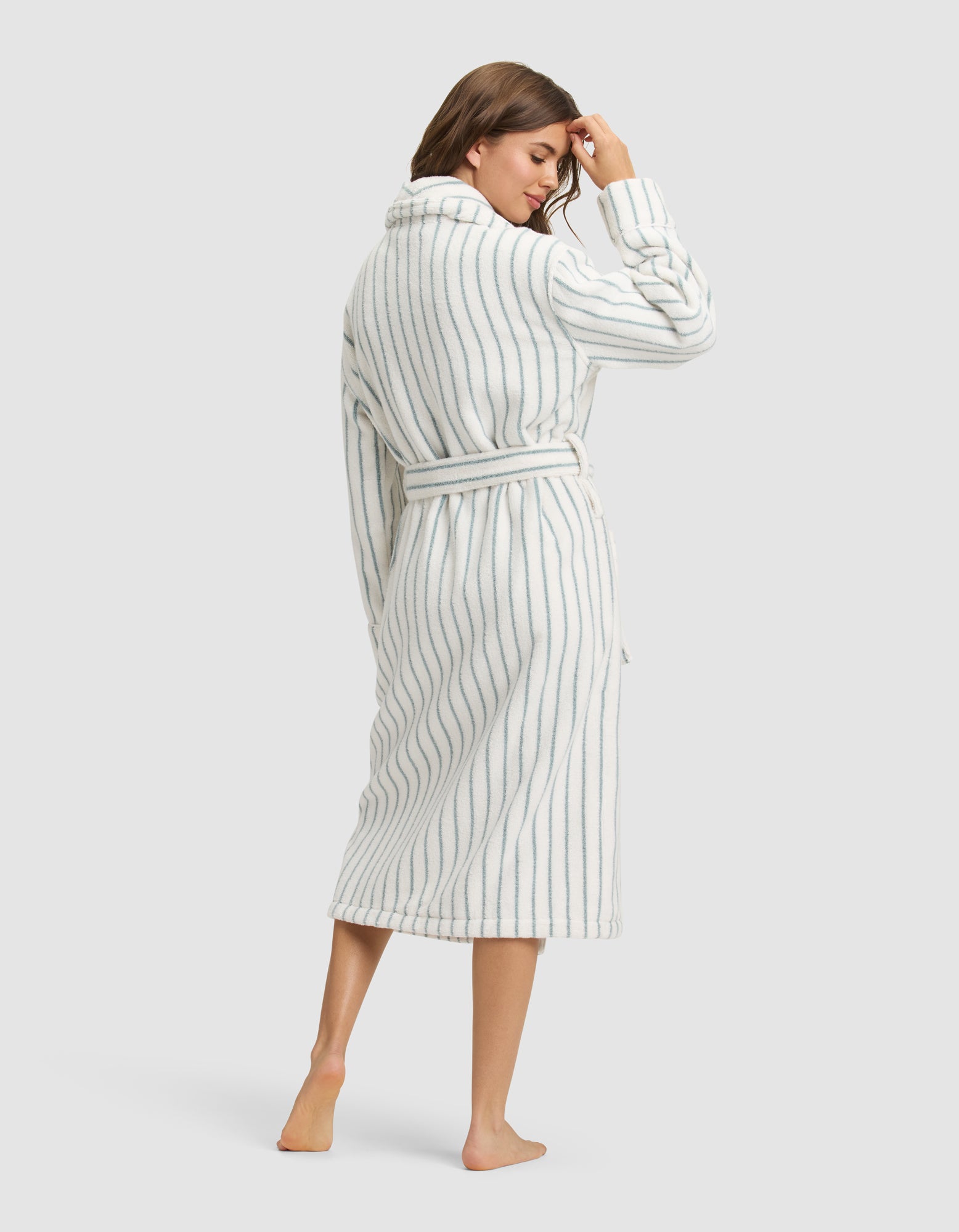 A woman stands barefoot, facing away, wearing the Cozy Earth Striped Luxe Bath Robe—a long, belted white robe with vertical blue stripes—touching her hair against a plain light background. |Color:Brook Stripe