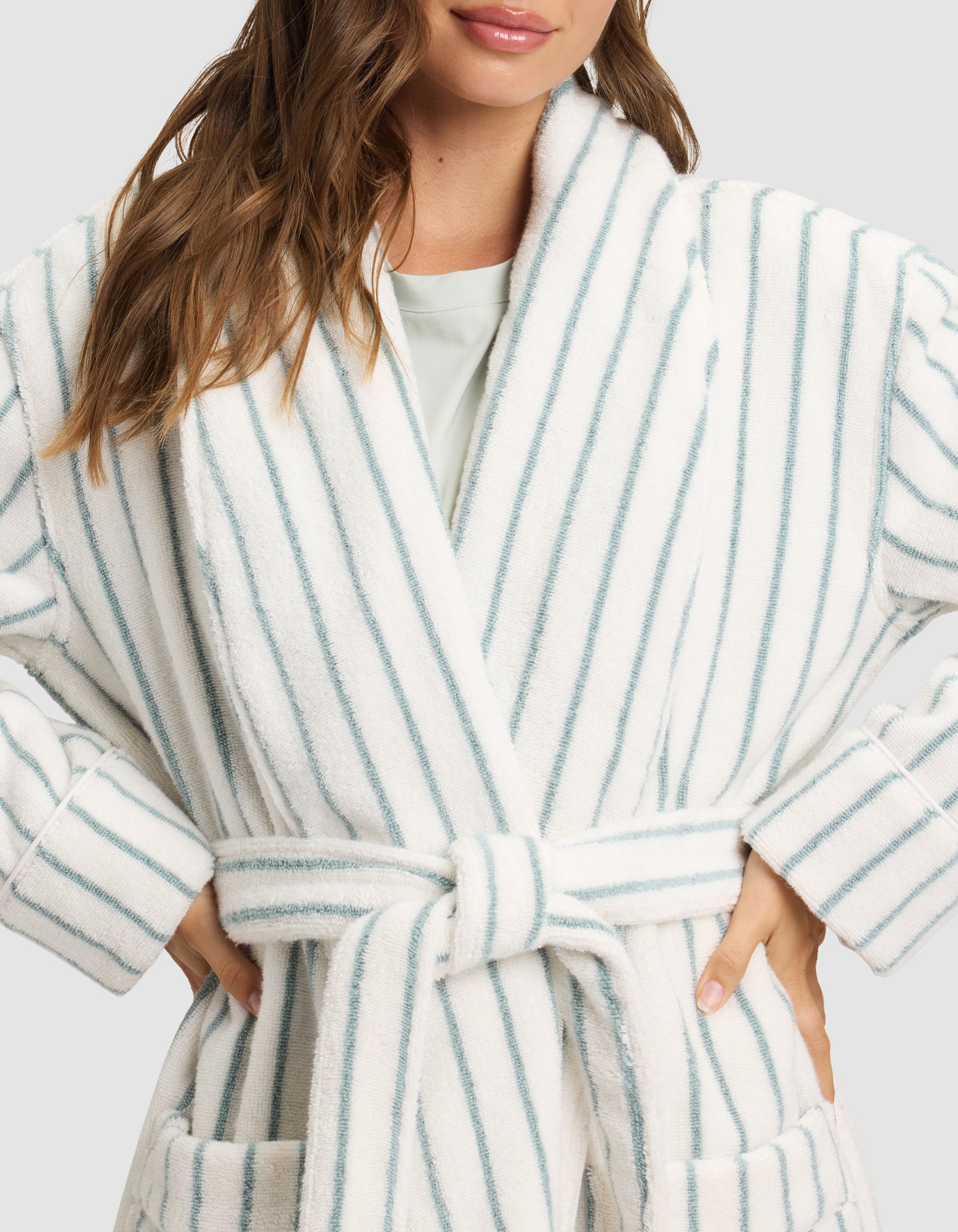 A woman models the Cozy Earth Striped Luxe Bath Robe, featuring white fabric with light blue vertical stripes, a belted waist, and large front pockets. Only her lower face and upper body are shown. 