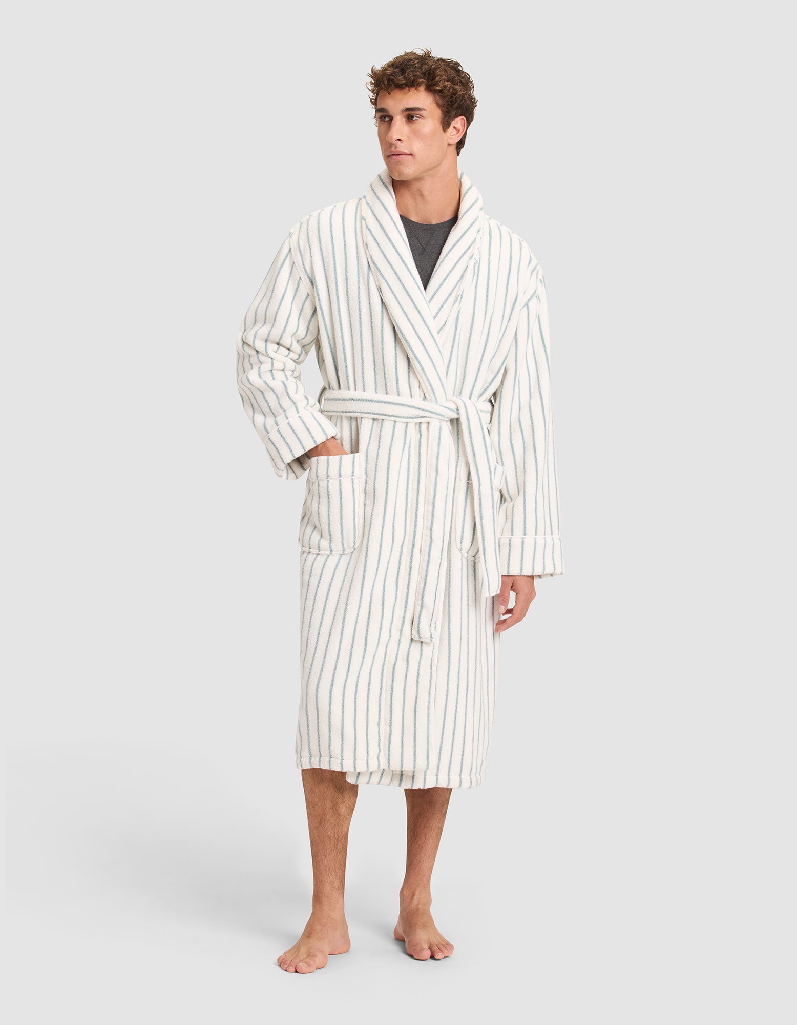 A man models the Cozy Earth Striped Luxe Bath Robe—white with thin gray stripes, tied at the waist over a dark shirt. He stands barefoot with one hand in his pocket, set against a plain light background. 