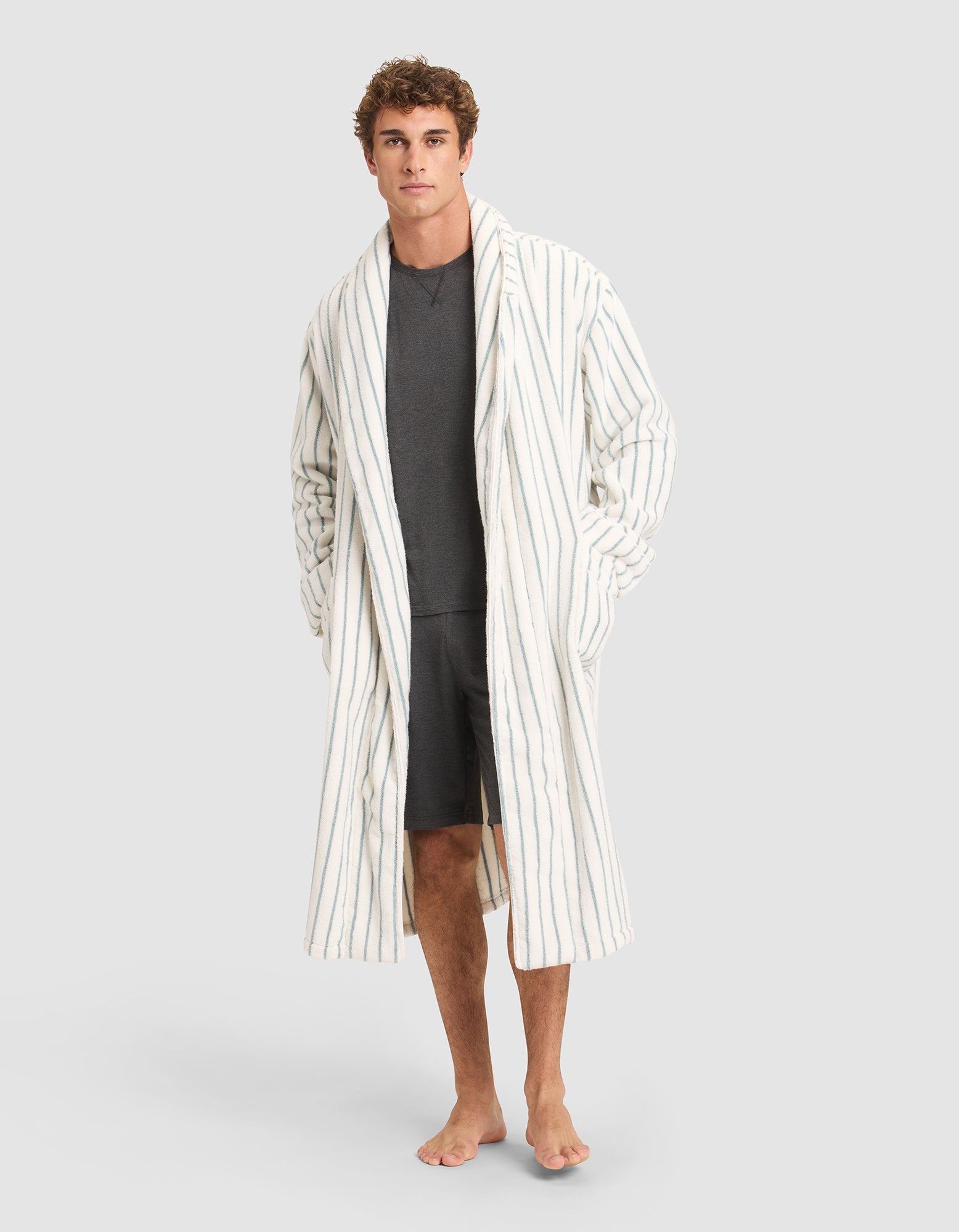 A man stands barefoot in a dark gray shirt and shorts, wearing the Cozy Earth Striped Luxe Bath Robe—a long, white robe with thin vertical blue stripes—against a plain light gray background. 