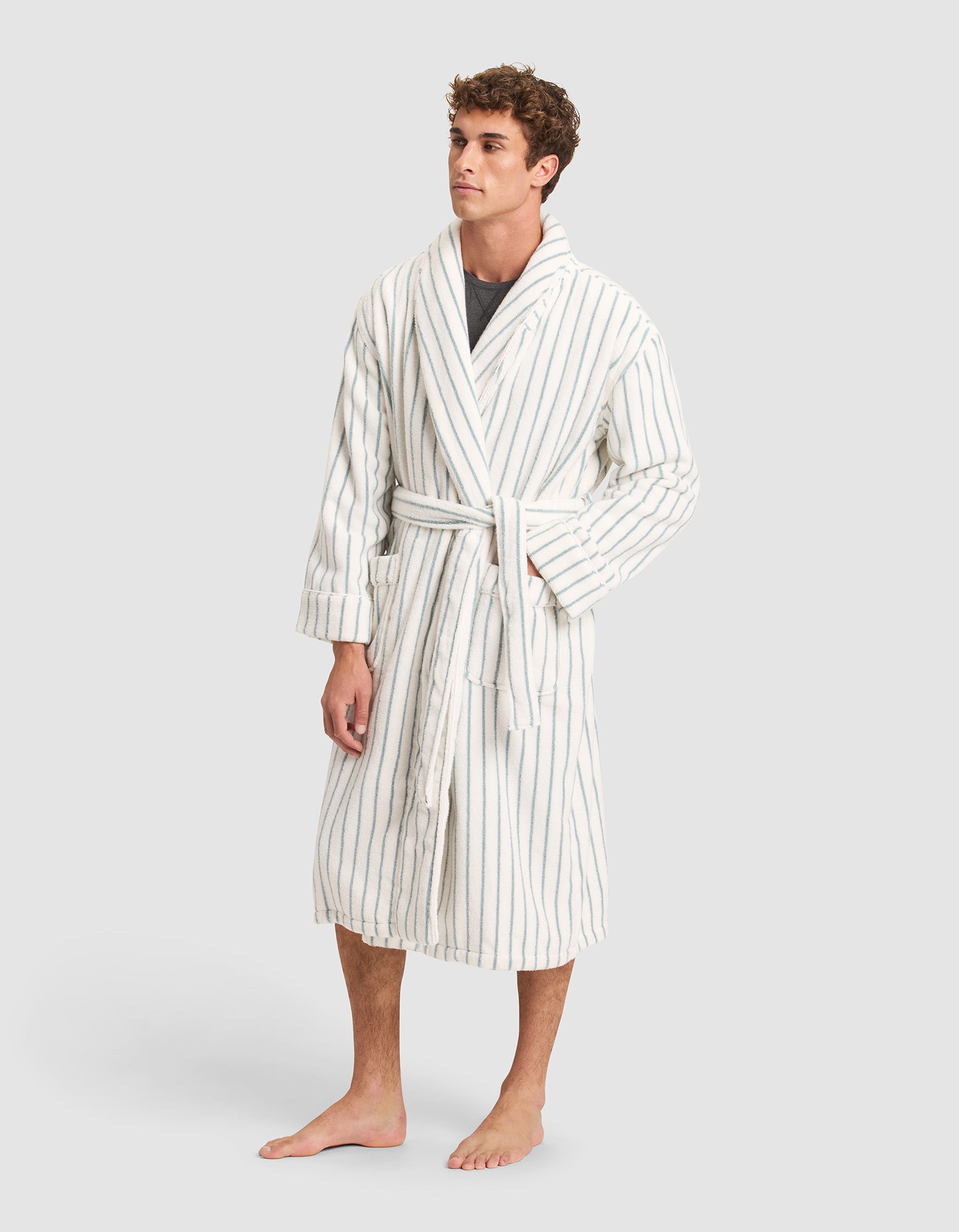 A man models the Cozy Earth Striped Luxe Bath Robe, featuring thin blue vertical stripes on white, standing barefoot against a plain light background and looking slightly to the side. 