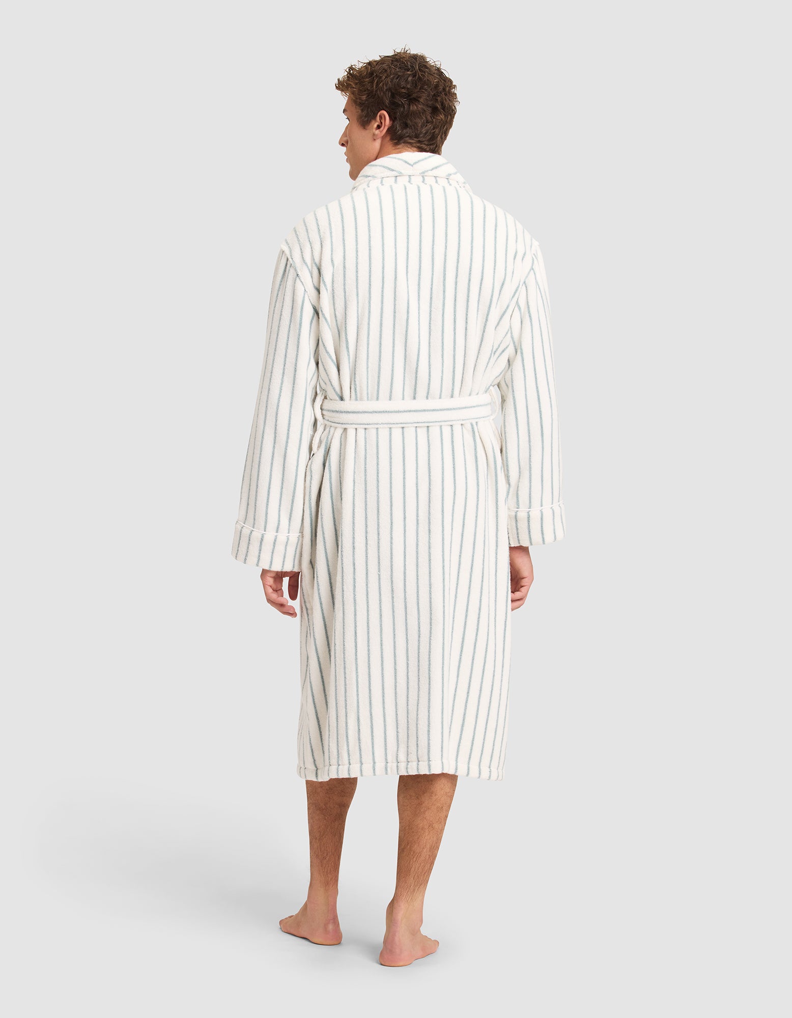 A man stands barefoot, facing away, wearing the Striped Luxe Bath Robe by Cozy Earth—white with vertical gray stripes and a belt—set against a plain light gray background. 