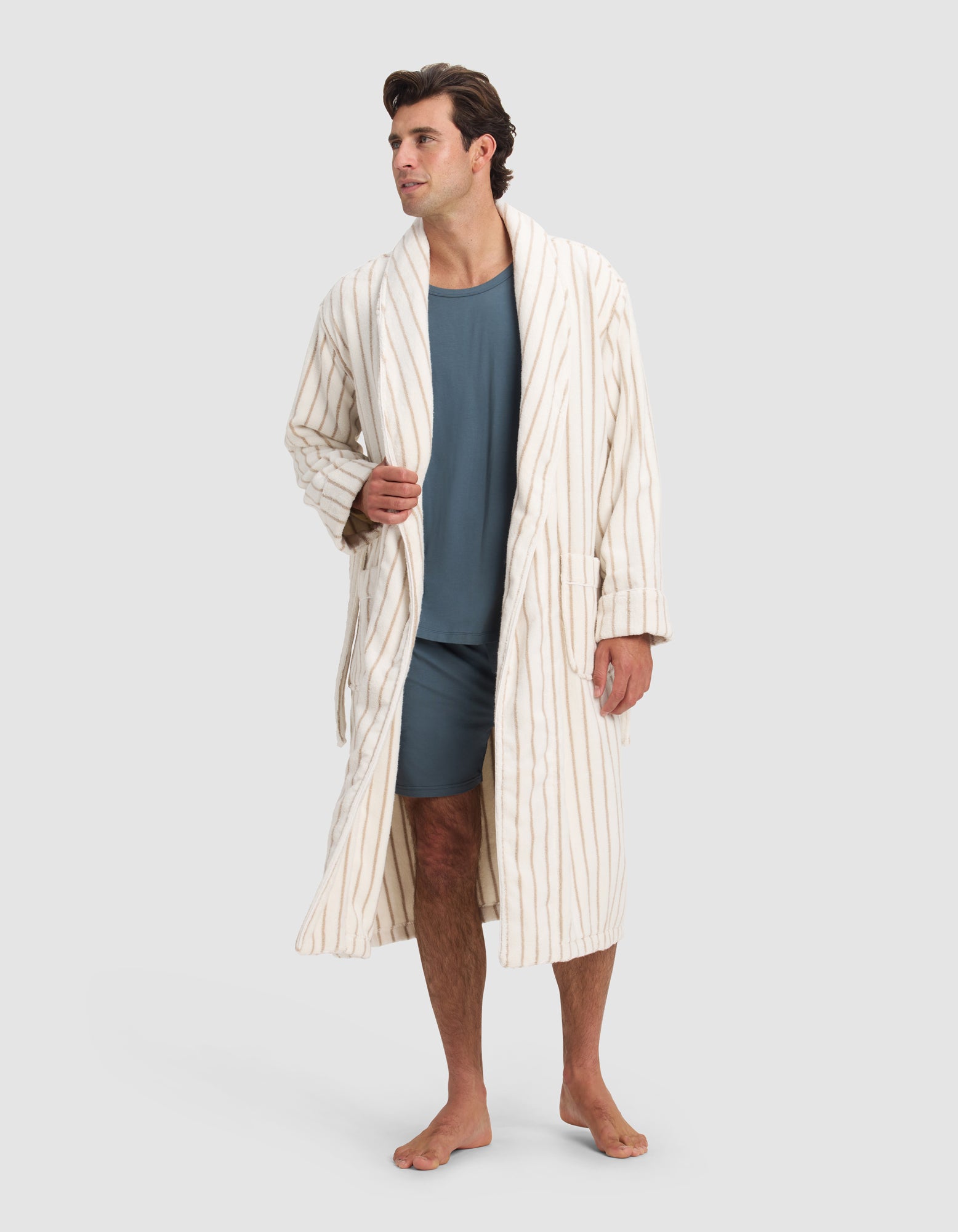A man stands barefoot in the Luxe Bath Robe by Cozy Earth—a long, cream robe with brown vertical stripes—over a blue t-shirt and shorts, looking to the side against a plain light background.
