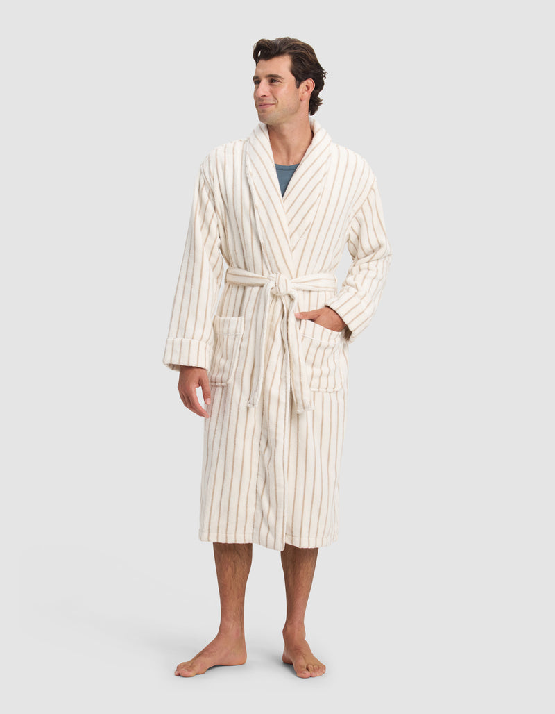 A man stands barefoot, smiling slightly and looking to the side, wearing Cozy Earth's Luxe Bath Robe in cream with thin beige stripes, a belted waist, two front pockets, and a gray shirt underneath.