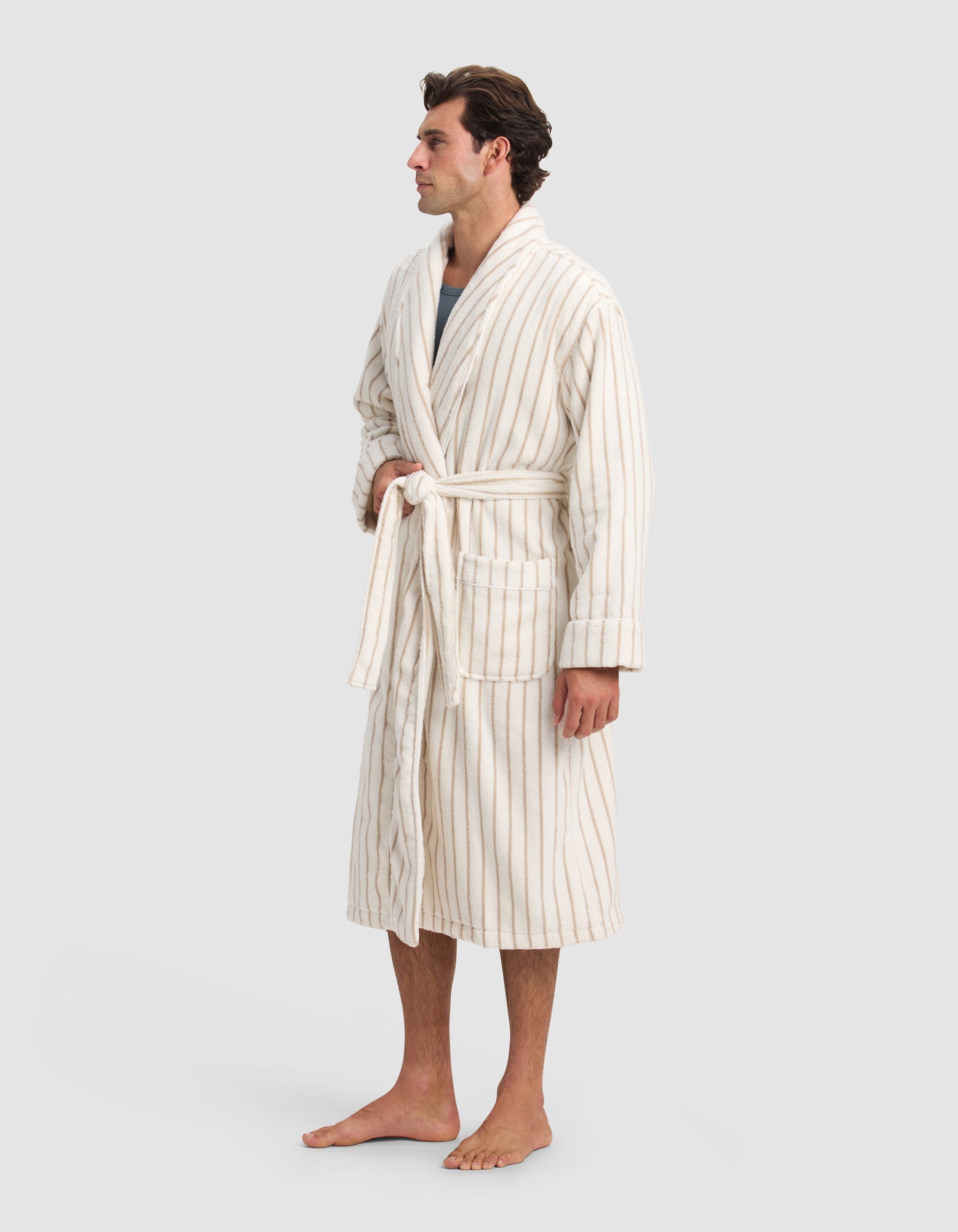 A man stands barefoot against a plain light gray background, wearing the Cozy Earth Luxe Bath Robe—a cream-colored, mid-length robe with thin vertical beige stripes and a tied belt.