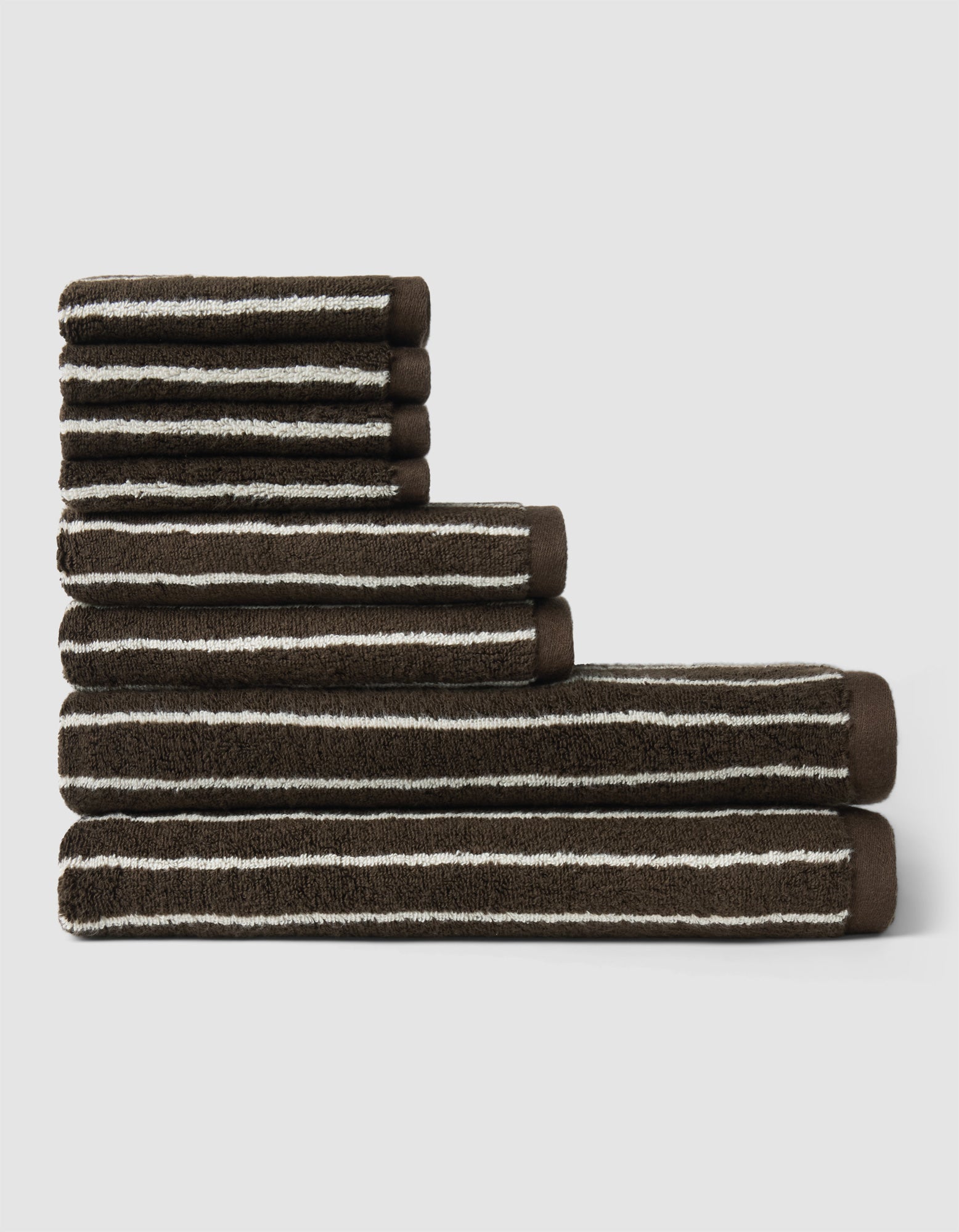 The Cozy Earth Striped Luxe Bath Towel Set, featuring dark brown towels with thin white stripes, is arranged in descending size order against a plain light gray background.