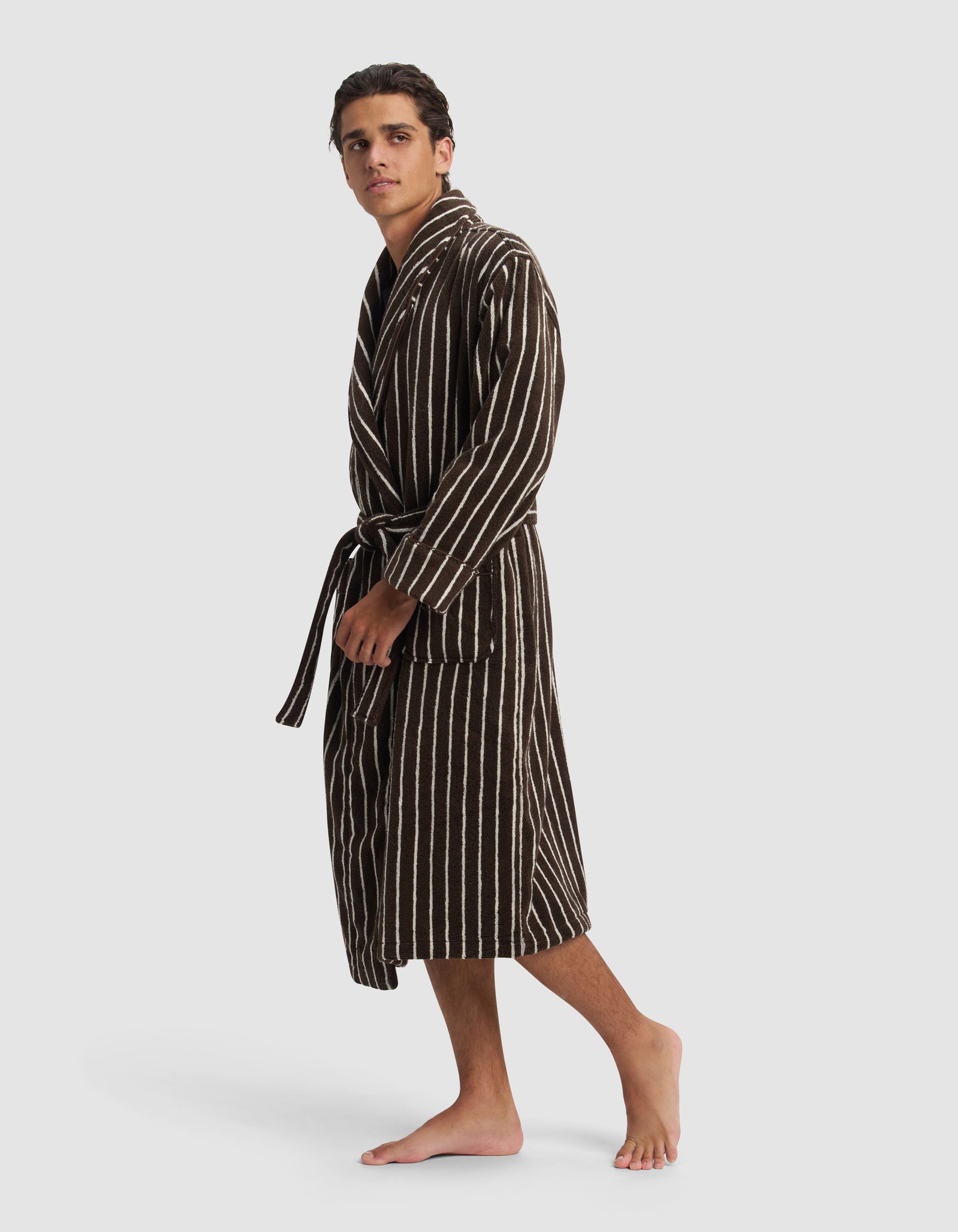A young man stands barefoot against a white background, wearing Cozy Earth's Striped Luxe Bath Robe in dark brown with vertical white stripes and a waist tie, gazing slightly to the side.