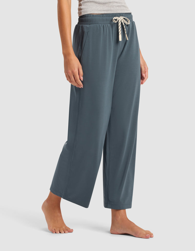 A woman wears Cozy Earth's Women's Studio Cropped Wide Leg Pant—loose, blue-gray with an elastic waistband and cream drawstring—standing barefoot against a plain light background.
