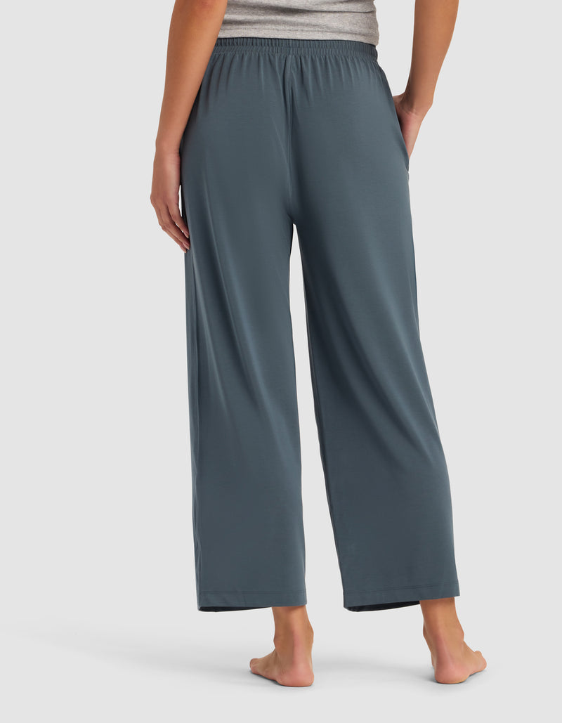 A woman stands barefoot, facing away, against a plain light background wearing Cozy Earth's Women's Studio Cropped Wide Leg Pant in blue-gray and a gray top.