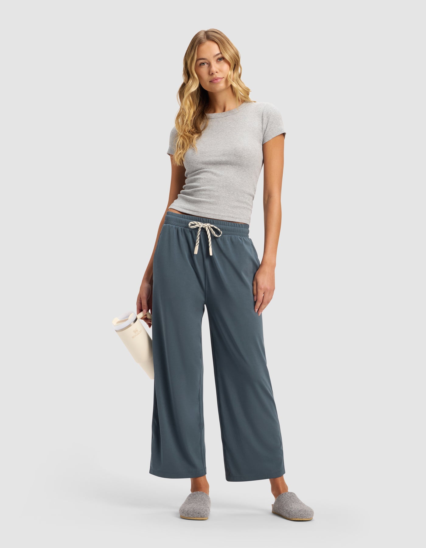 A woman with long blonde hair wears a fitted gray t-shirt, gray slippers, and Cozy Earth Women's Studio Cropped Wide Leg Pant in blue-gray. She holds a white water bottle and stands against a plain light background.