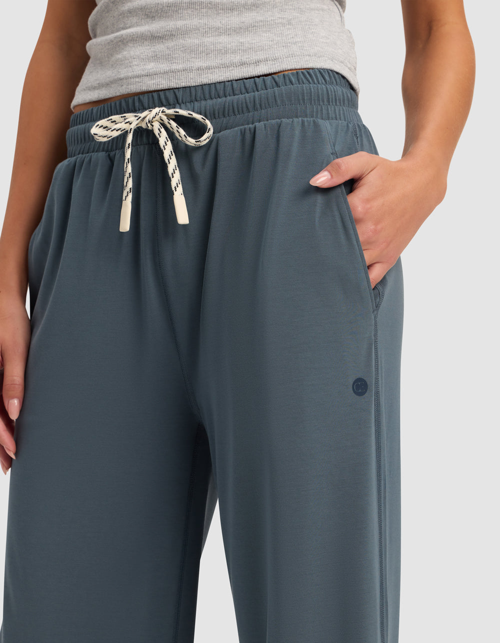A person in Cozy Earth's Women's Studio Cropped Wide Leg Pant—loose blue-gray joggers with a cream and black drawstring—pairs them with a gray ribbed tank top and stands with one hand in a pocket.