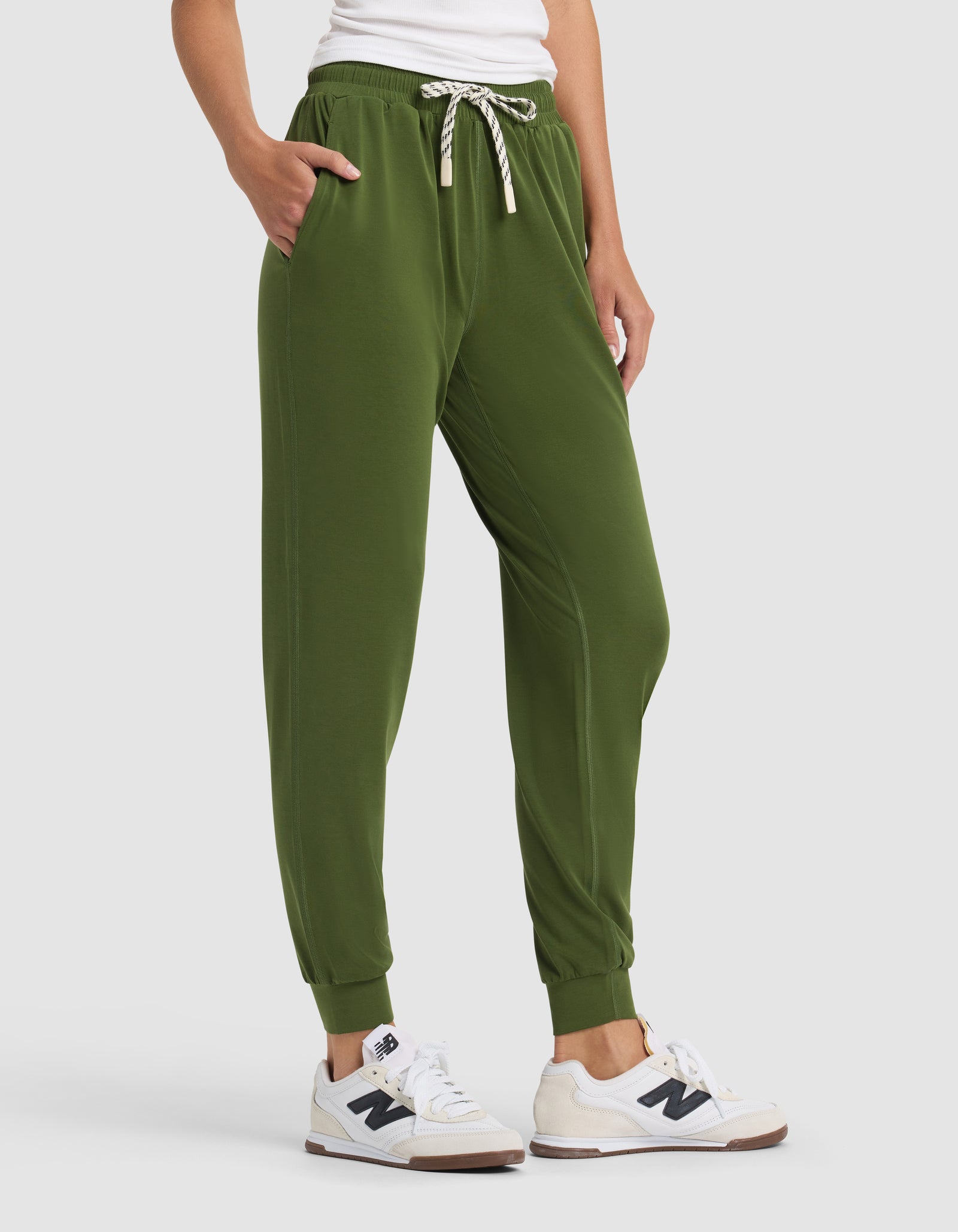 A person wears Cozy Earth's Women's Studio Jogger in olive green with a drawstring waist, paired with a white T-shirt and white sneakers, posing with one hand in their pocket against a plain background.