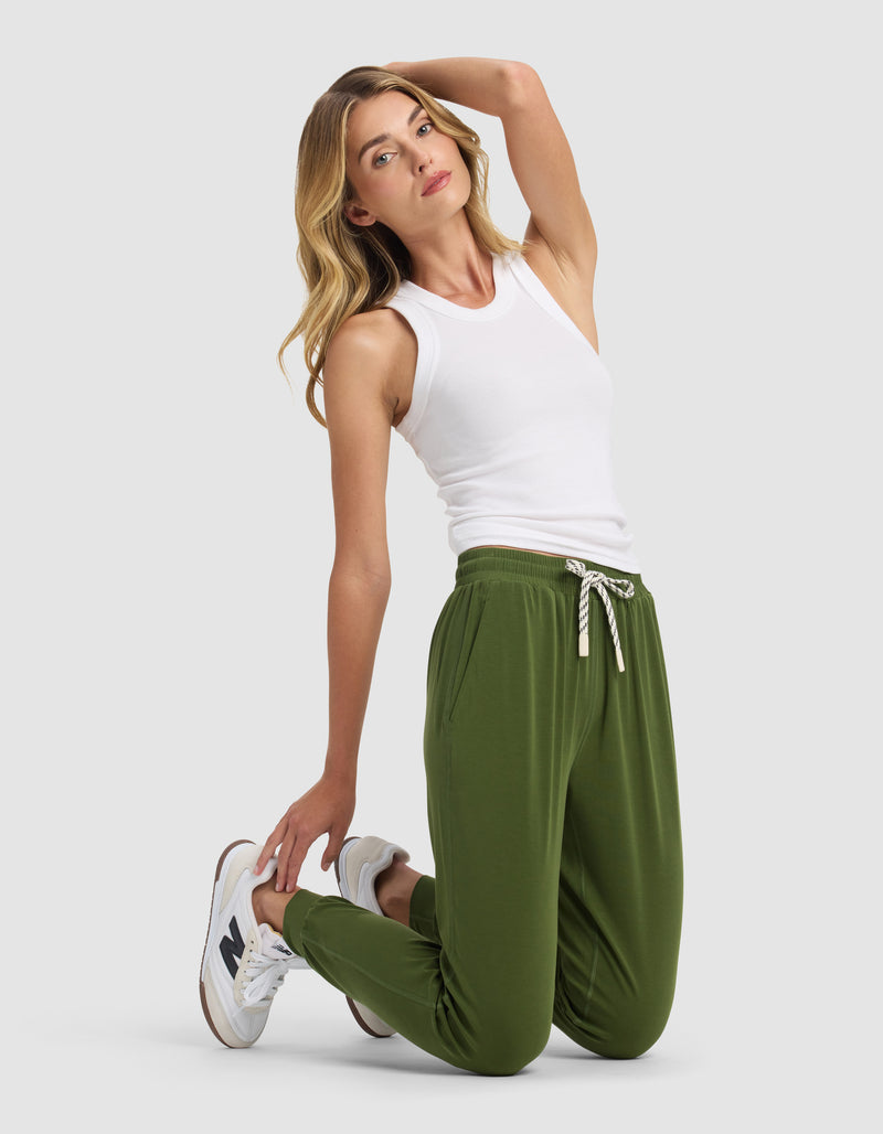 A woman with long, wavy blonde hair kneels on a light gray background, wearing Cozy Earth's Women's Studio Jogger in green, a white sleeveless top, and white sneakers. She poses with one hand behind her head.