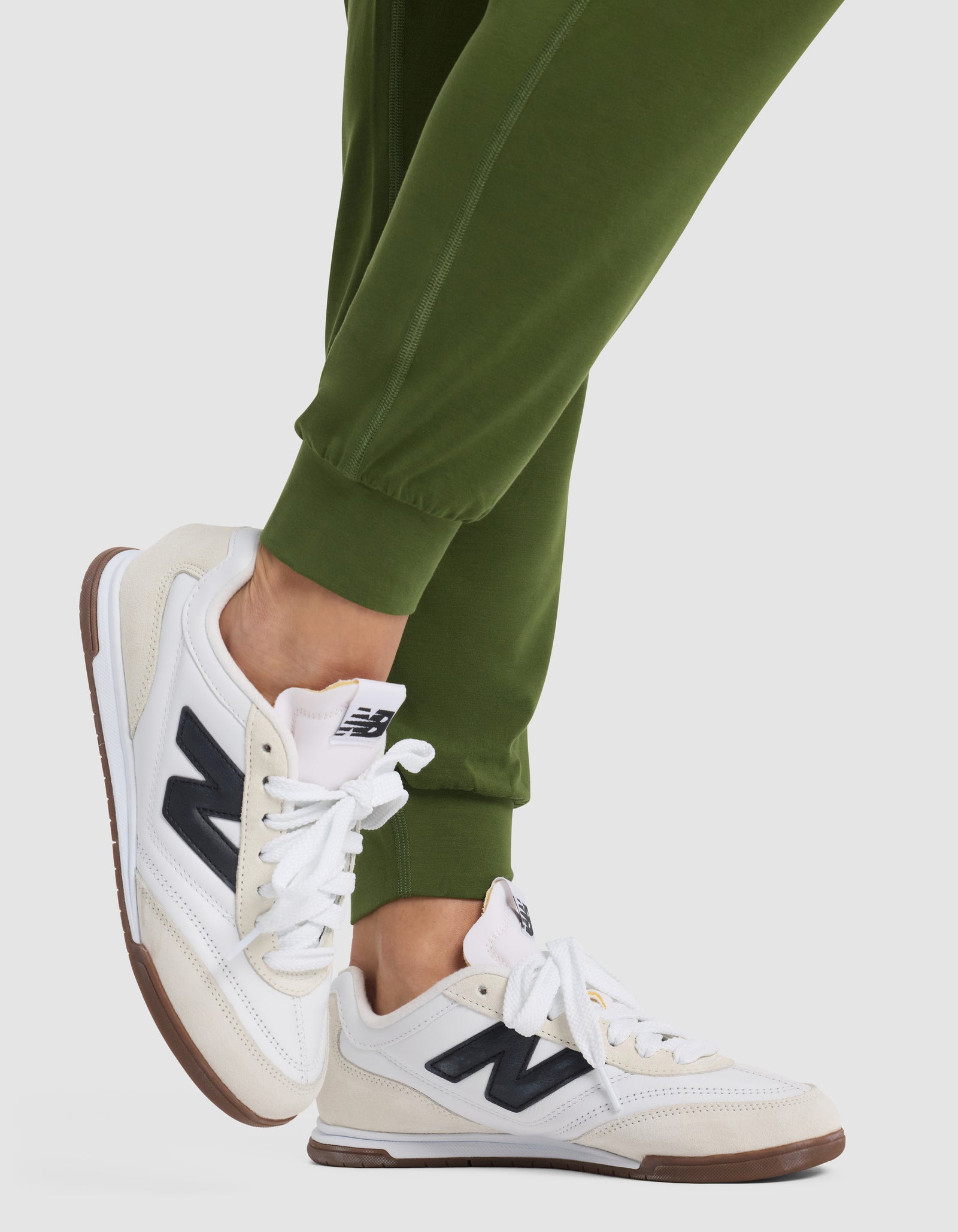 A person wearing Cozy Earth Women's Studio Jogger in green and white New Balance sneakers with black "N" logos stands on tiptoes against a plain light background.