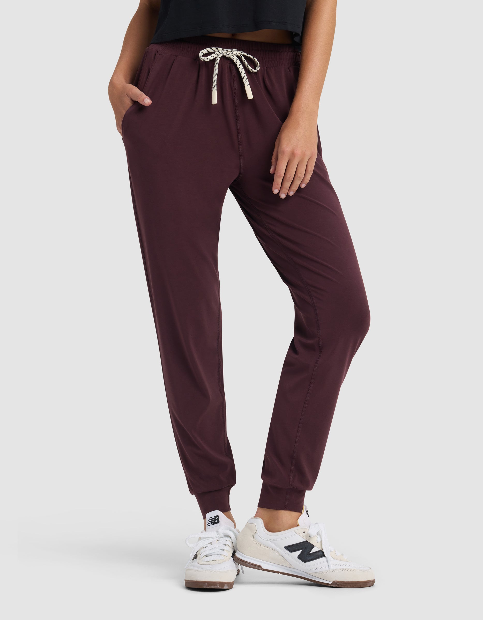 A person stands with hands in pockets against a plain light background, wearing Cozy Earth Women's Studio Jogger pants in maroon, a black top, and white New Balance sneakers.|Color:Raisin