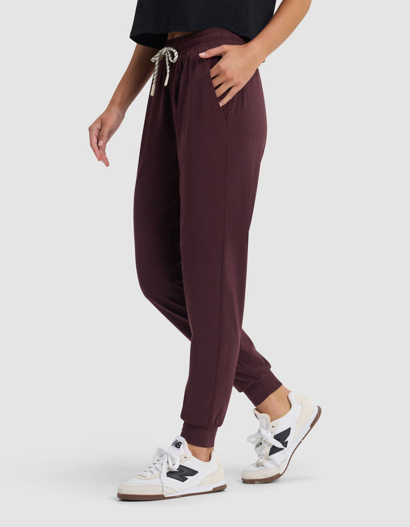 A person models the Cozy Earth Women's Studio Jogger in burgundy, paired with a black top and white sneakers with black accents, standing with one hand in a pocket against a plain light background.