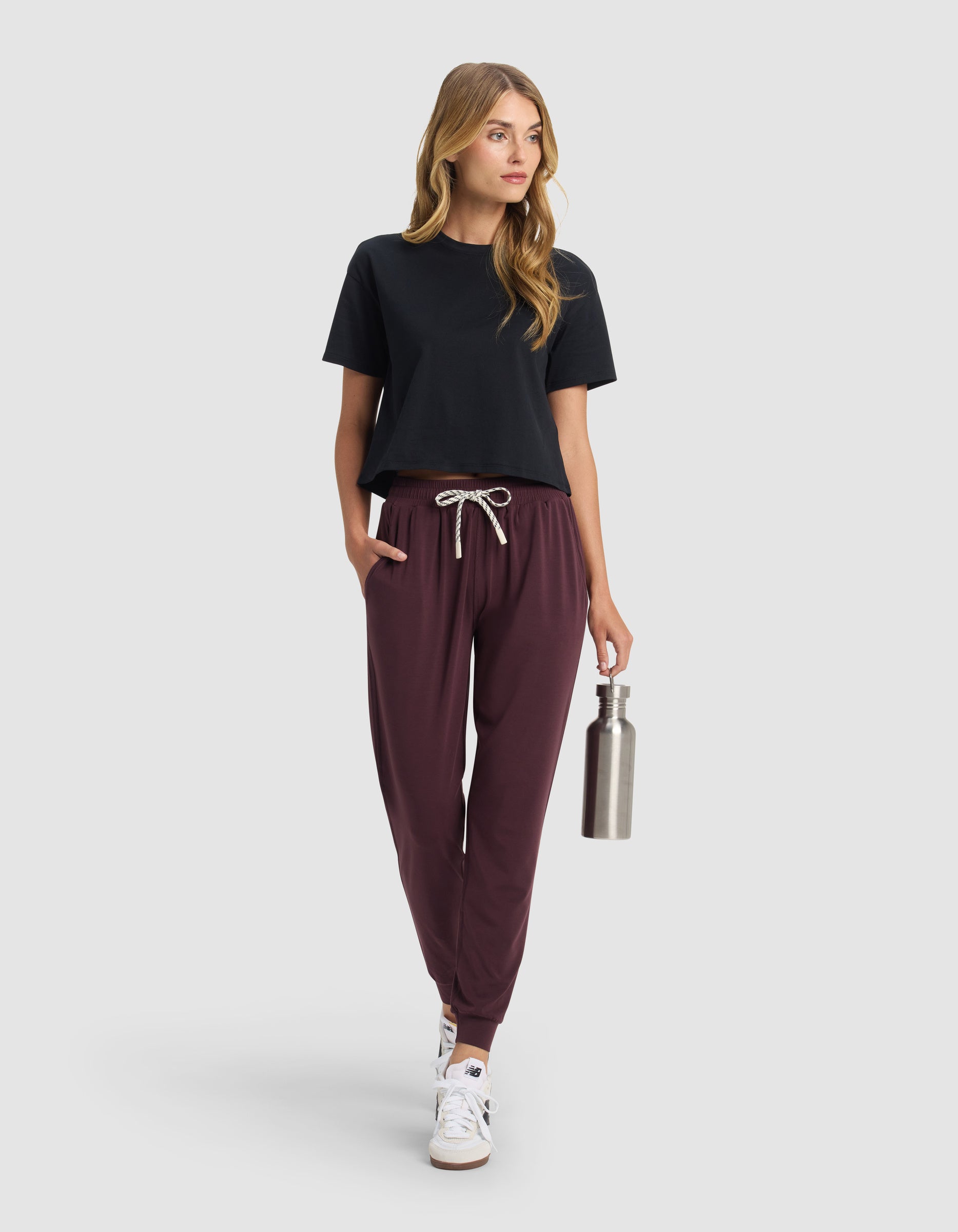 A woman with long hair models Cozy Earth's Women's Studio Jogger in maroon, paired with a black t-shirt and white sneakers. She holds a stainless steel water bottle and looks to the side against a plain light gray background.