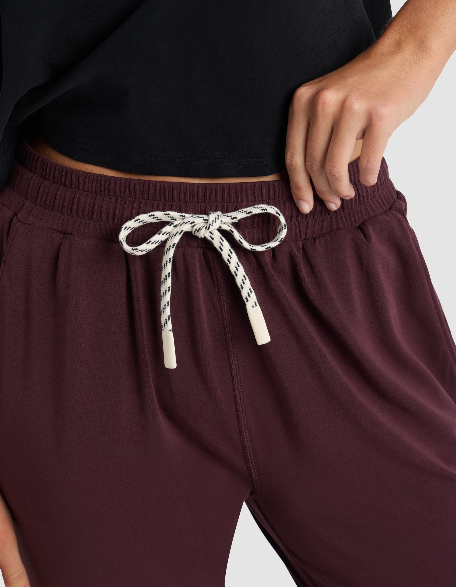 A woman models the Cozy Earth Women's Studio Jogger in maroon, featuring a white and black patterned drawstring tied in a bow, paired with a black top and posing with one hand on her hip.
