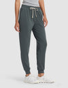 A person wearing Cozy Earth's Women's Studio Jogger in dark green, paired with a light gray top and white athletic shoes, stands against a plain light background.|Color:Storm