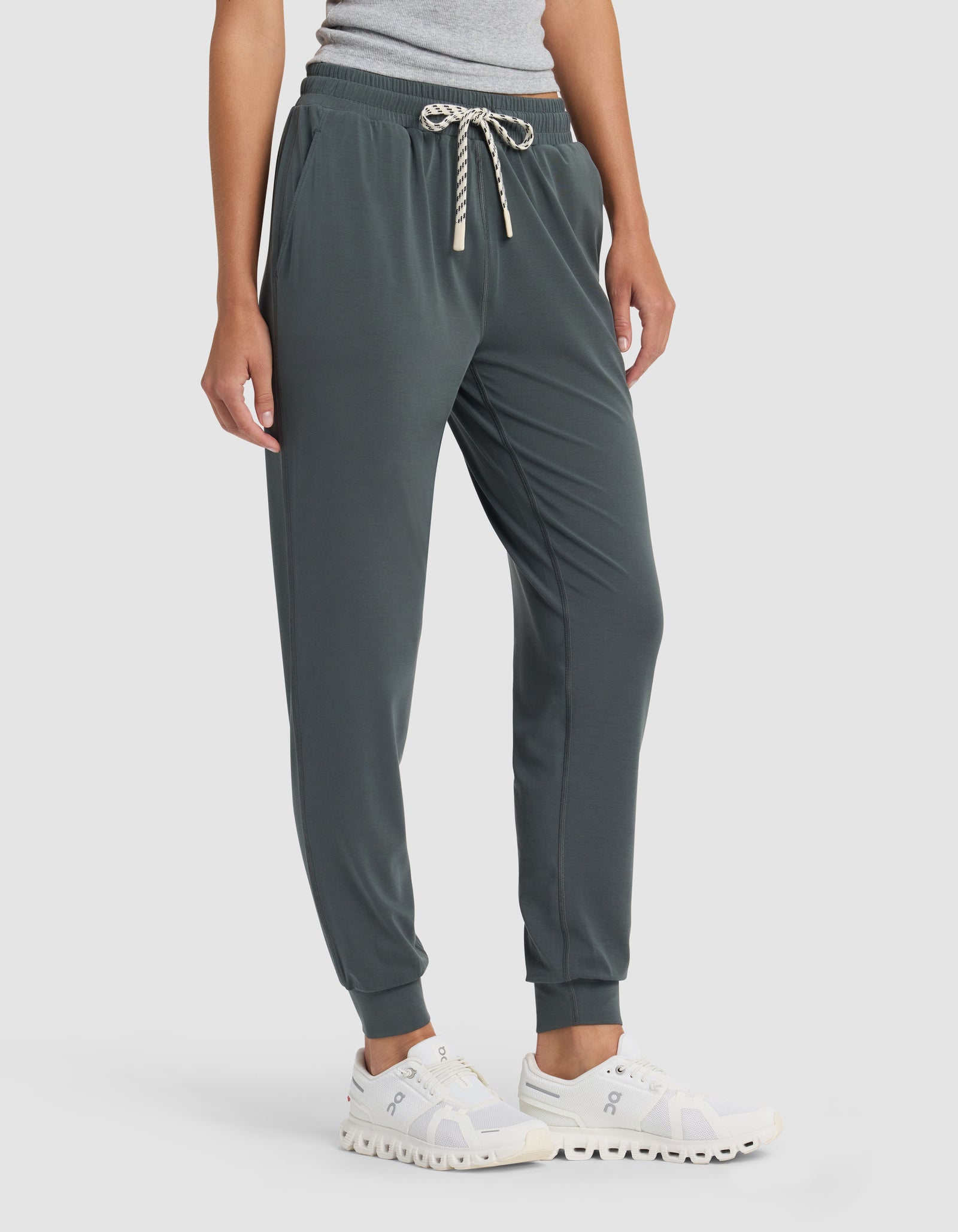 A person wearing Cozy Earth's Women's Studio Jogger in dark green, paired with a light gray top and white athletic shoes, stands against a plain light background.