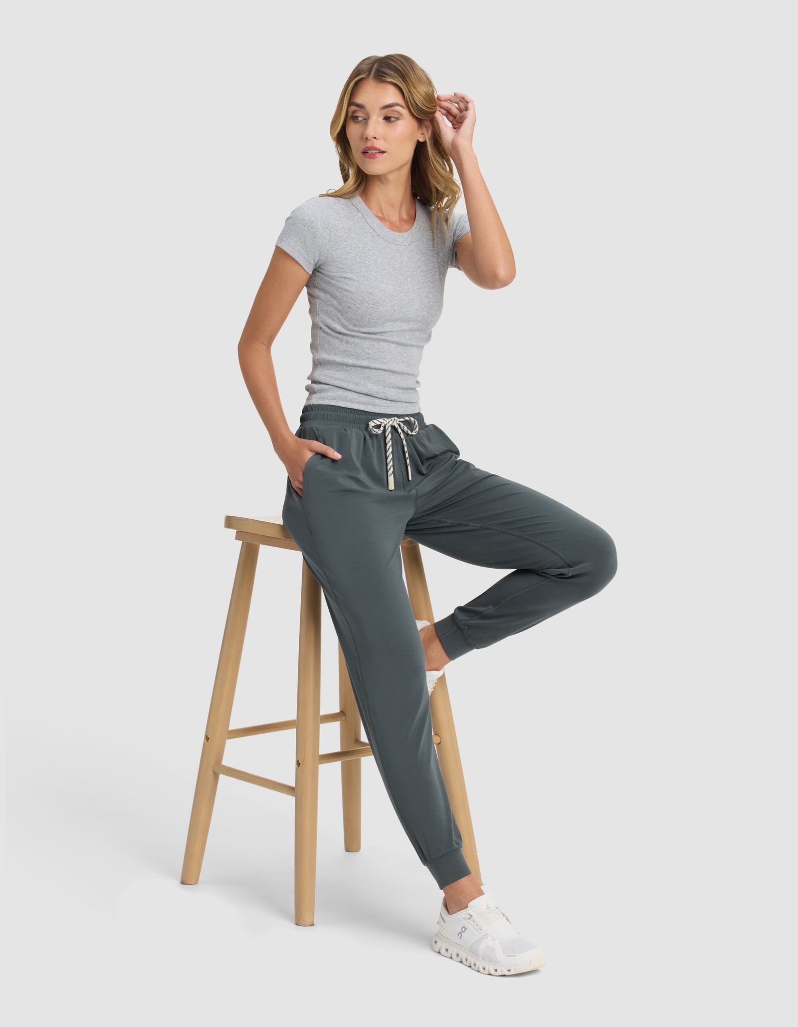 A woman in Cozy Earth Women's Studio Jogger and a fitted gray t-shirt sits on a wooden stool against a light background, wearing white sneakers, one hand in her pocket and the other touching her hair.