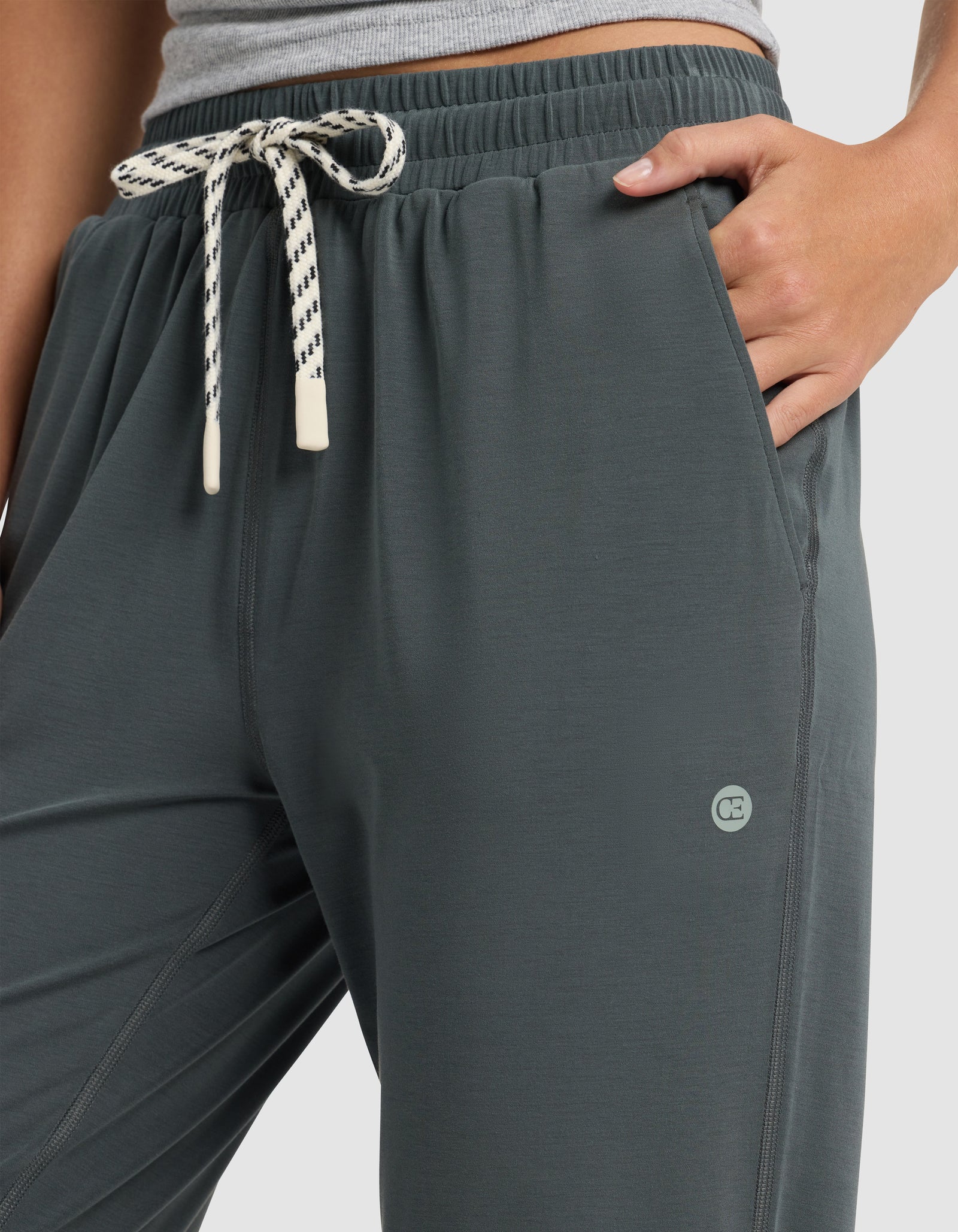 A person wears Cozy Earth's Women's Studio Jogger in dark grey, featuring a white and black drawstring, small circular logo by the left pocket, paired with a light grey top and one hand in the jogger's pocket.