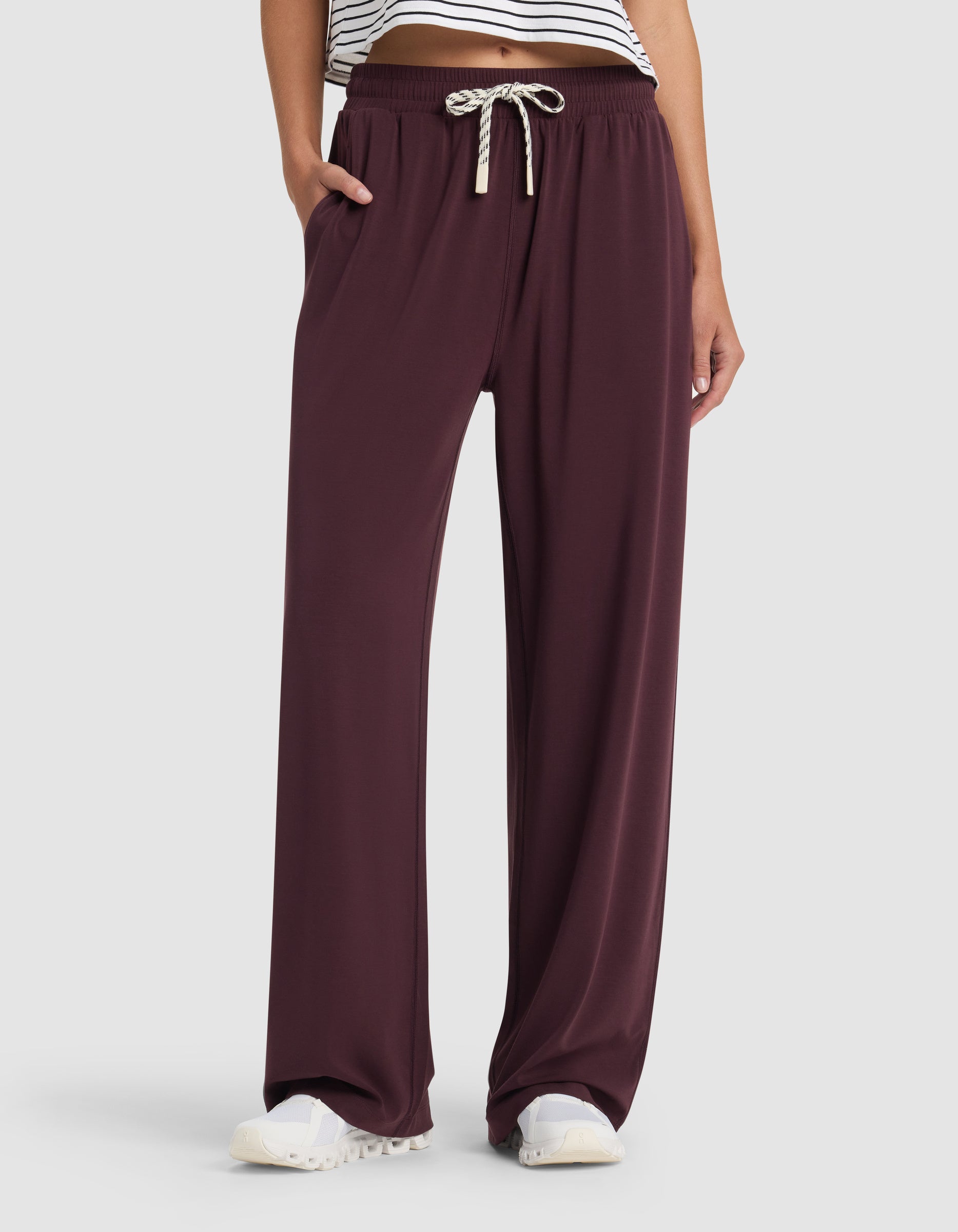 A person wears Cozy Earth Women's Studio Wide Leg Pant in burgundy with a white drawstring, white sneakers, and a cropped striped top. Their left hand is in their pocket; only the lower body is shown. |Color:Raisin