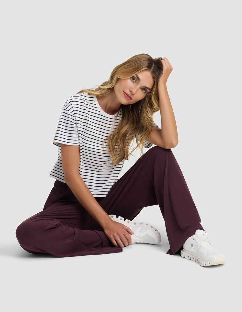 A woman with long blonde hair sits on the floor against a plain background, wearing Cozy Earth's Women's Studio Wide Leg Pant in maroon, paired with a black and white striped t-shirt and white sneakers. She rests her head on one hand, gazing at the camera.
