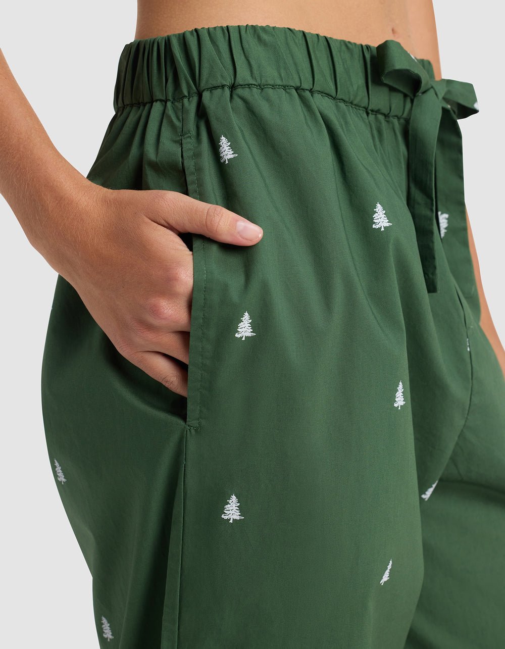 Close-up of a person wearing Cozy Earth's Women's Sutton Long Sleeve Pajama Set featuring green pants with white embroidered trees, an elastic waistband, and a fabric tie belt, their right hand resting in the pocket.