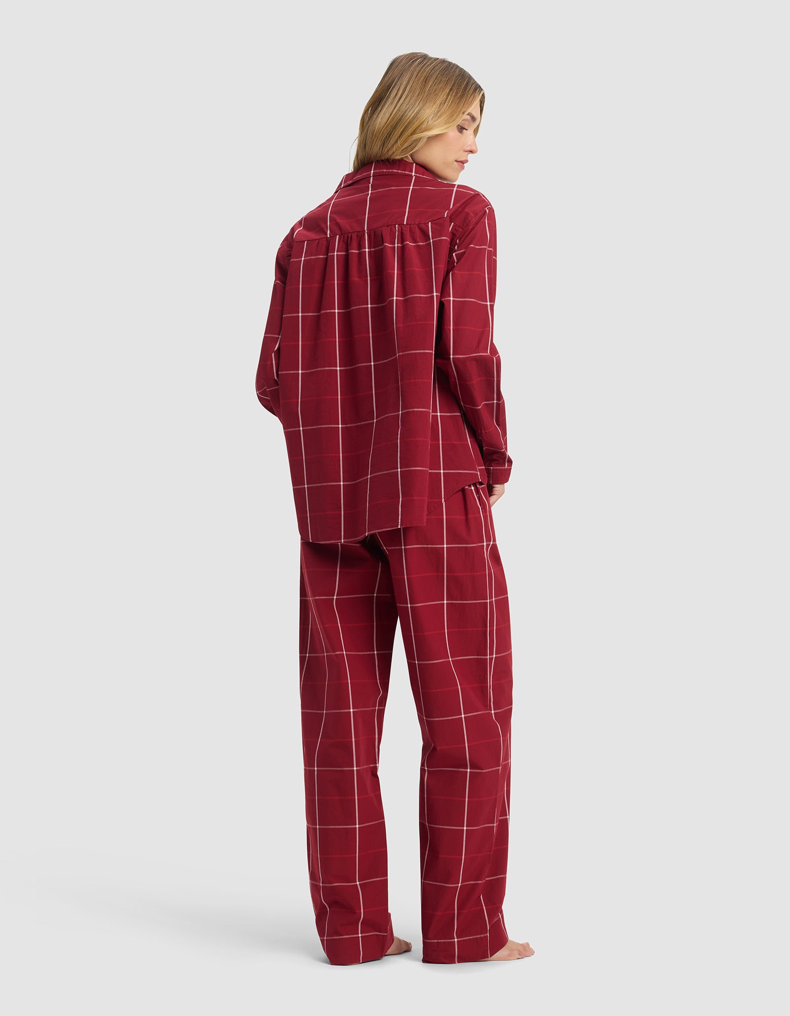 A woman faces away barefoot, wearing Cozy Earth's Women's Sutton Long Sleeve Pajama Set in red plaid with white grid lines. The relaxed-fit top and loose pants stand out against a plain light gray background.|Color:Garnet Windowpane