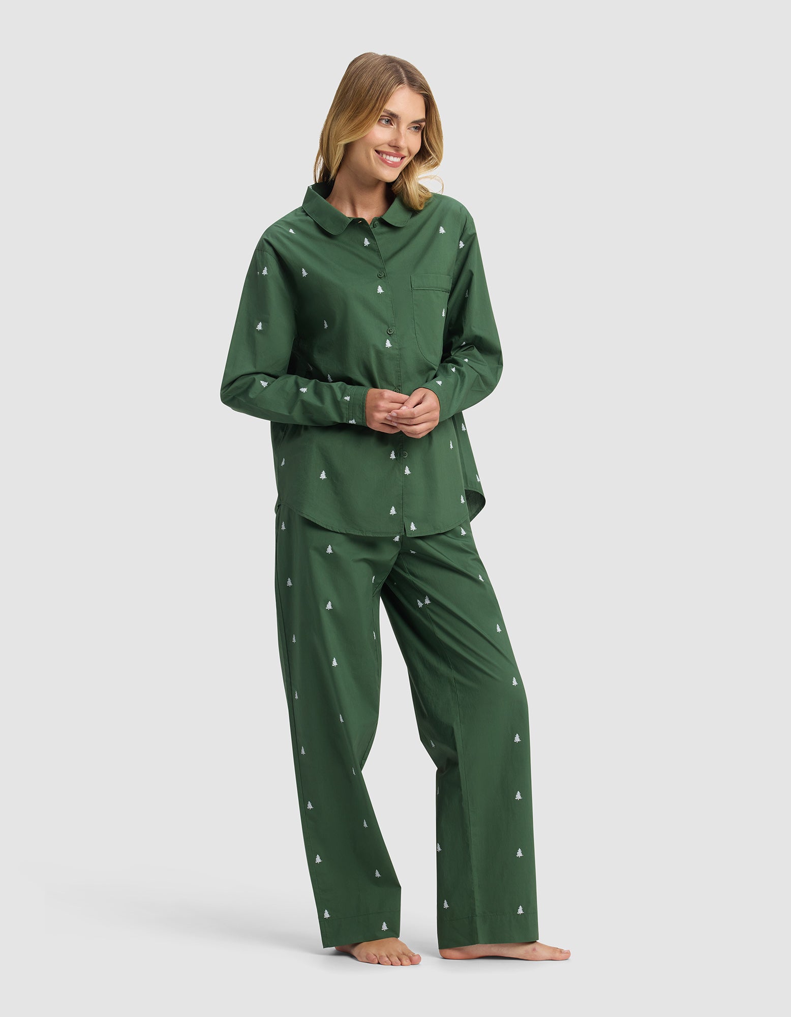 A woman stands barefoot and smiling against a plain light background, wearing Cozy Earth's Women's Sutton Long Sleeve Pajama Set in dark green with small white tree patterns.|Color:Jade Tree