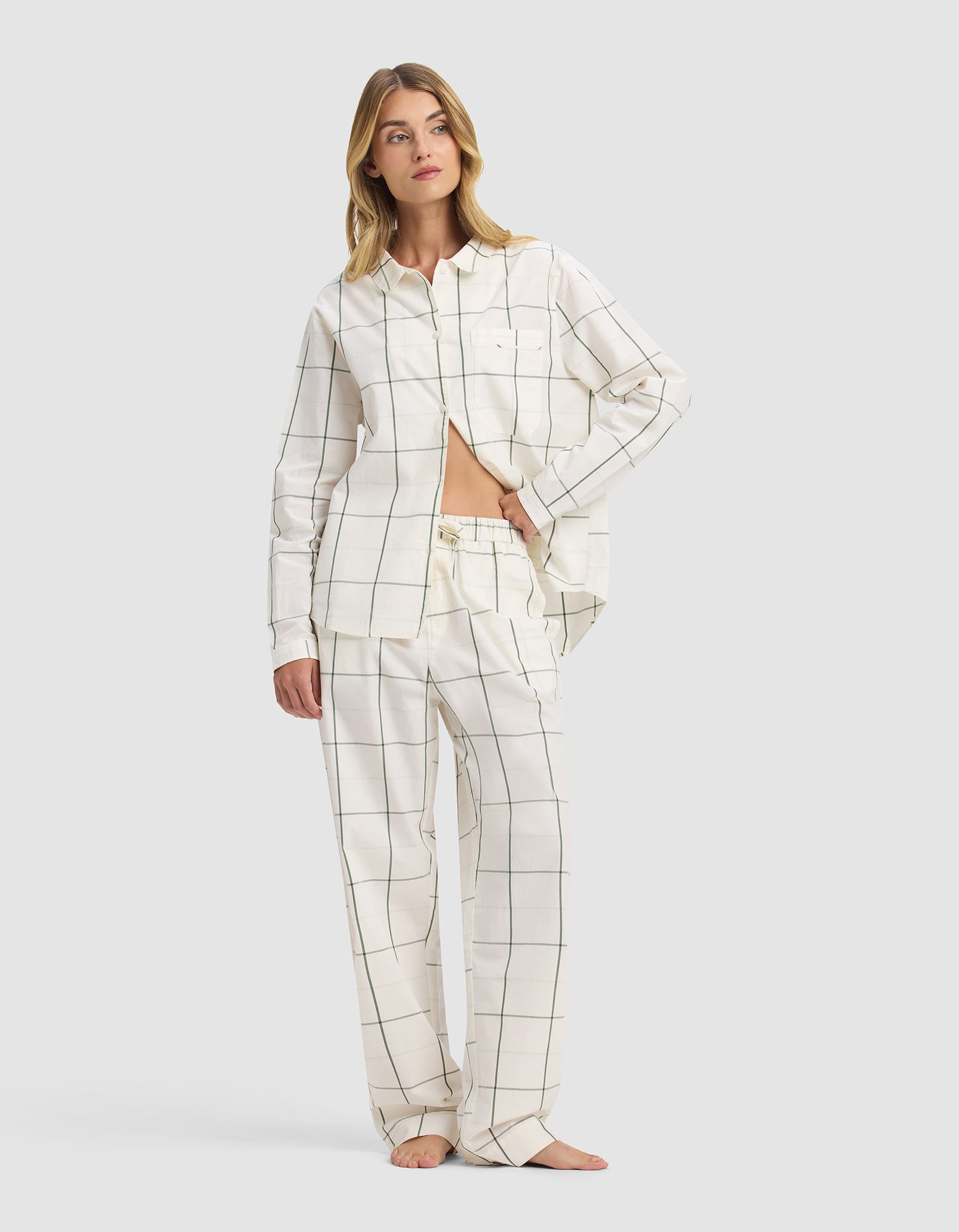 A woman stands barefoot wearing Cozy Earth's Sutton Pajama Pant and matching long-sleeve button-up, both in a white fabric with a minimalist black grid pattern. She poses with one hand in her pocket and looks to the side.