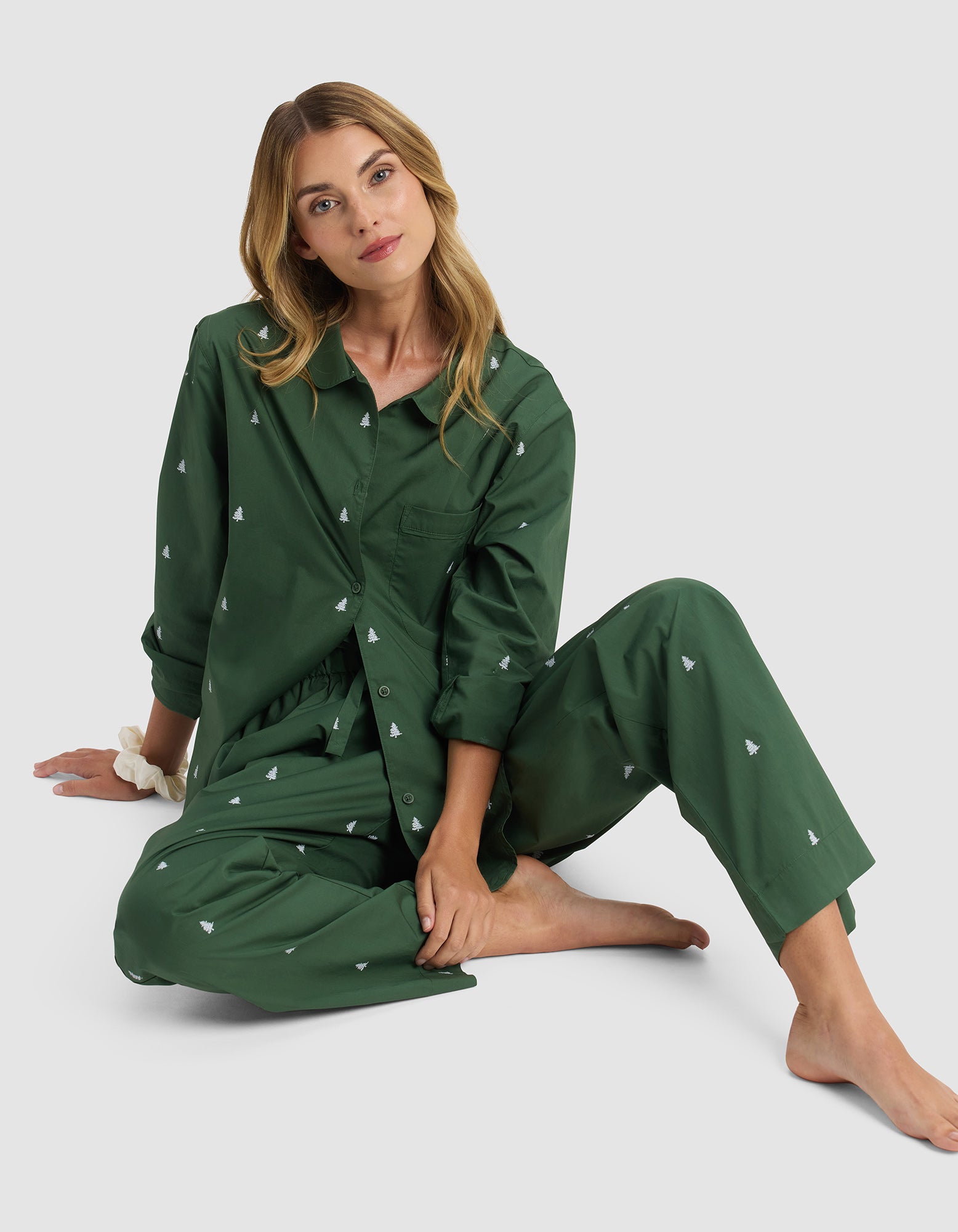 A woman with long blonde hair sits barefoot on the floor in Cozy Earth’s Sutton Pajama Pant, a matching dark green set featuring small white tree patterns, gazing calmly at the camera against a plain gray background.