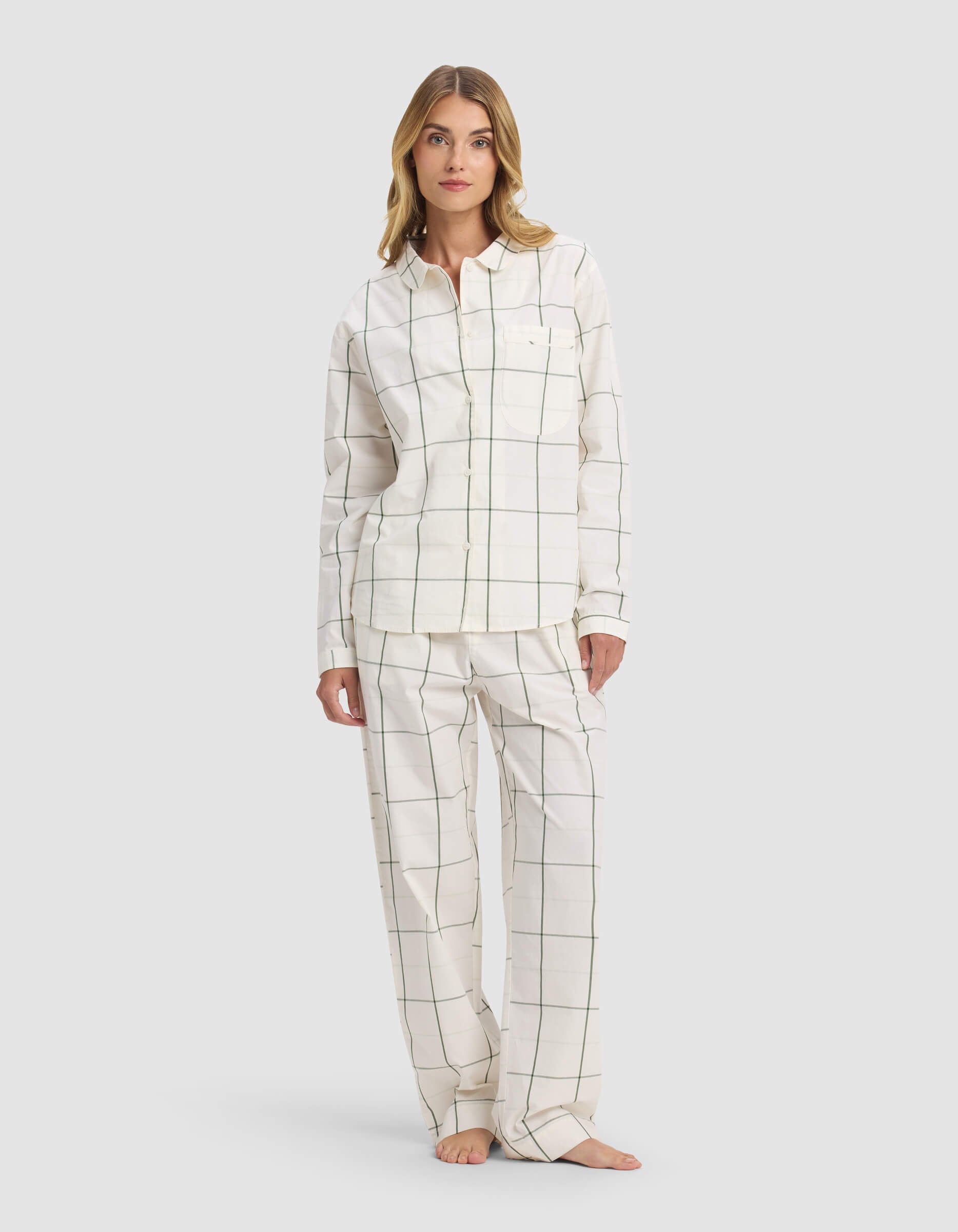 A woman models the Cozy Earth Women's Sutton Long Sleeve Pajama Set, featuring a white background with thin black grid lines, a button-up shirt, and loose pants. She stands barefoot facing forward with a neutral expression.