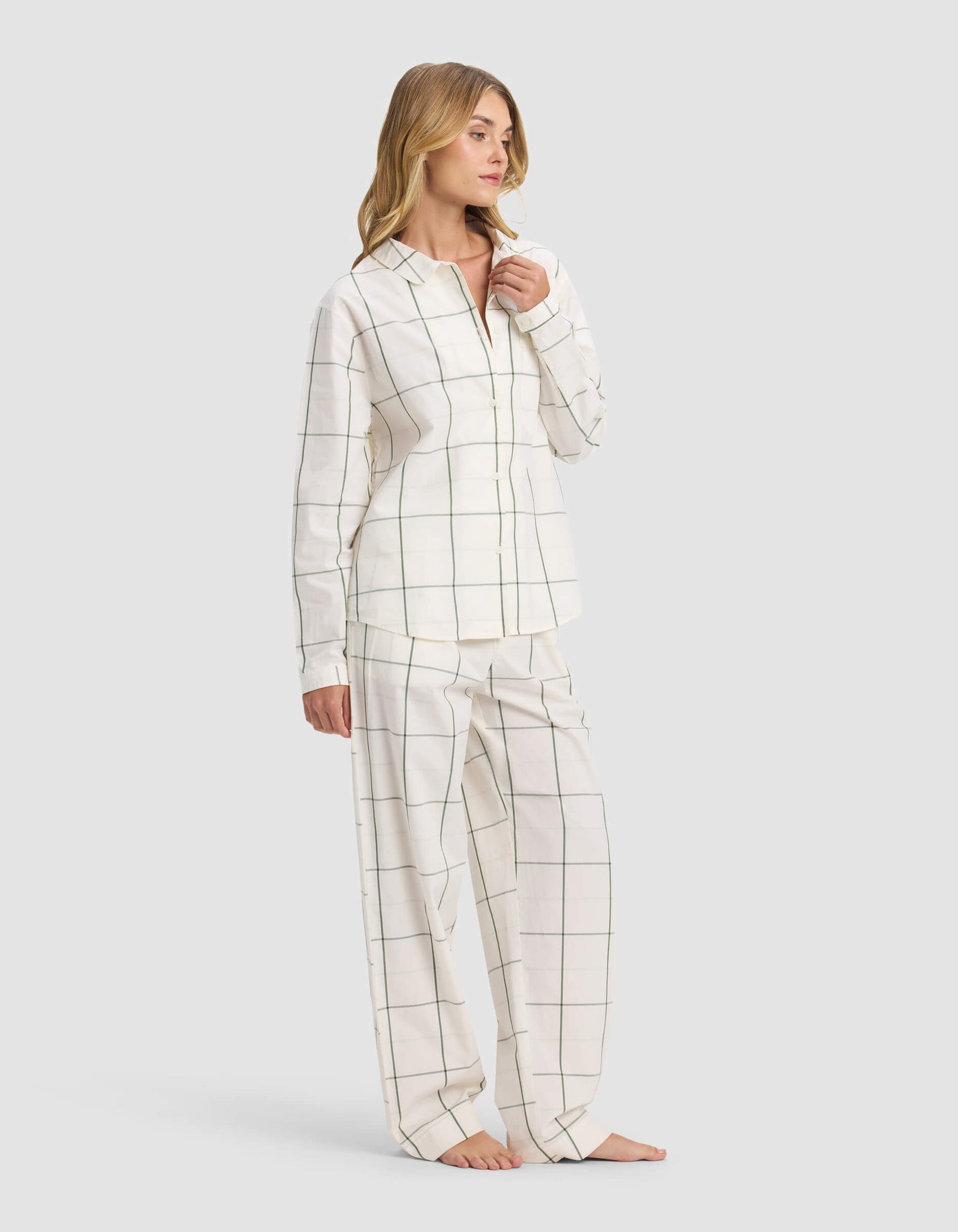 A woman stands barefoot, wearing the Cozy Earth Women's Sutton Long Sleeve Pajama Set in white with thin black grid lines, featuring a button-up shirt and loose pants. She looks to the side and touches her chin.