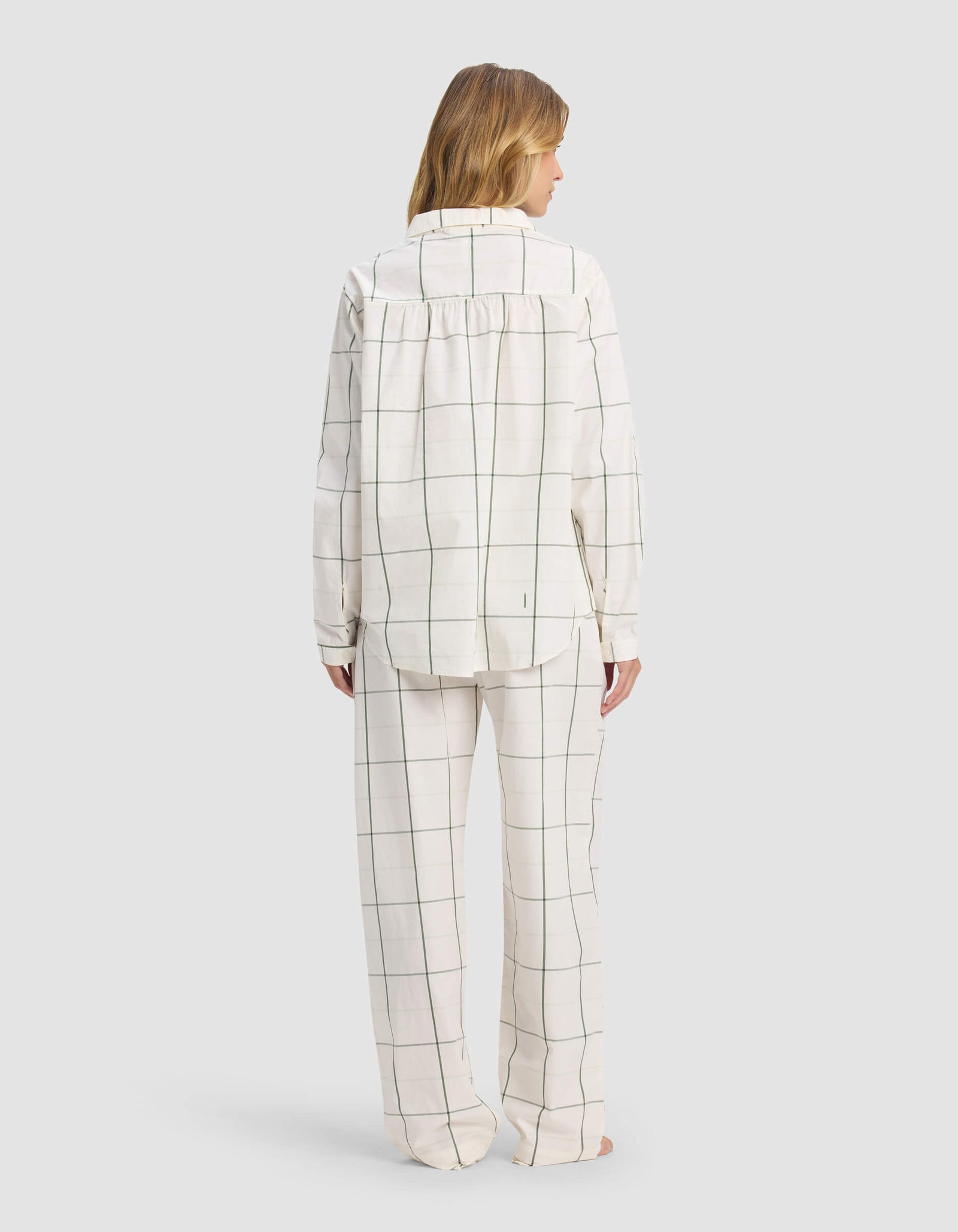 A person faces away, barefoot, in Cozy Earth’s Women's Sutton Long Sleeve Pajama Set: loose white pajamas with a black windowpane grid, collared top with long sleeves, straight-leg pants. The background is plain gray.