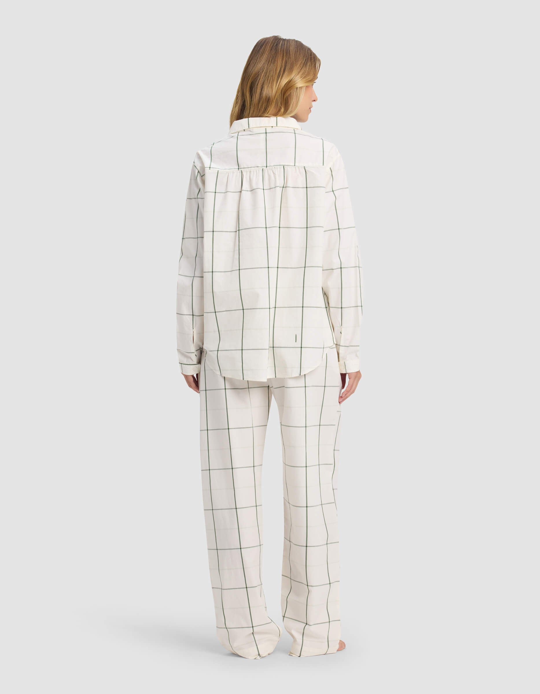 A person faces away, barefoot, in Cozy Earth’s Women's Sutton Long Sleeve Pajama Set: loose white pajamas with a black windowpane grid, collared top with long sleeves, straight-leg pants. The background is plain gray.|Color:Bone Windowpane
