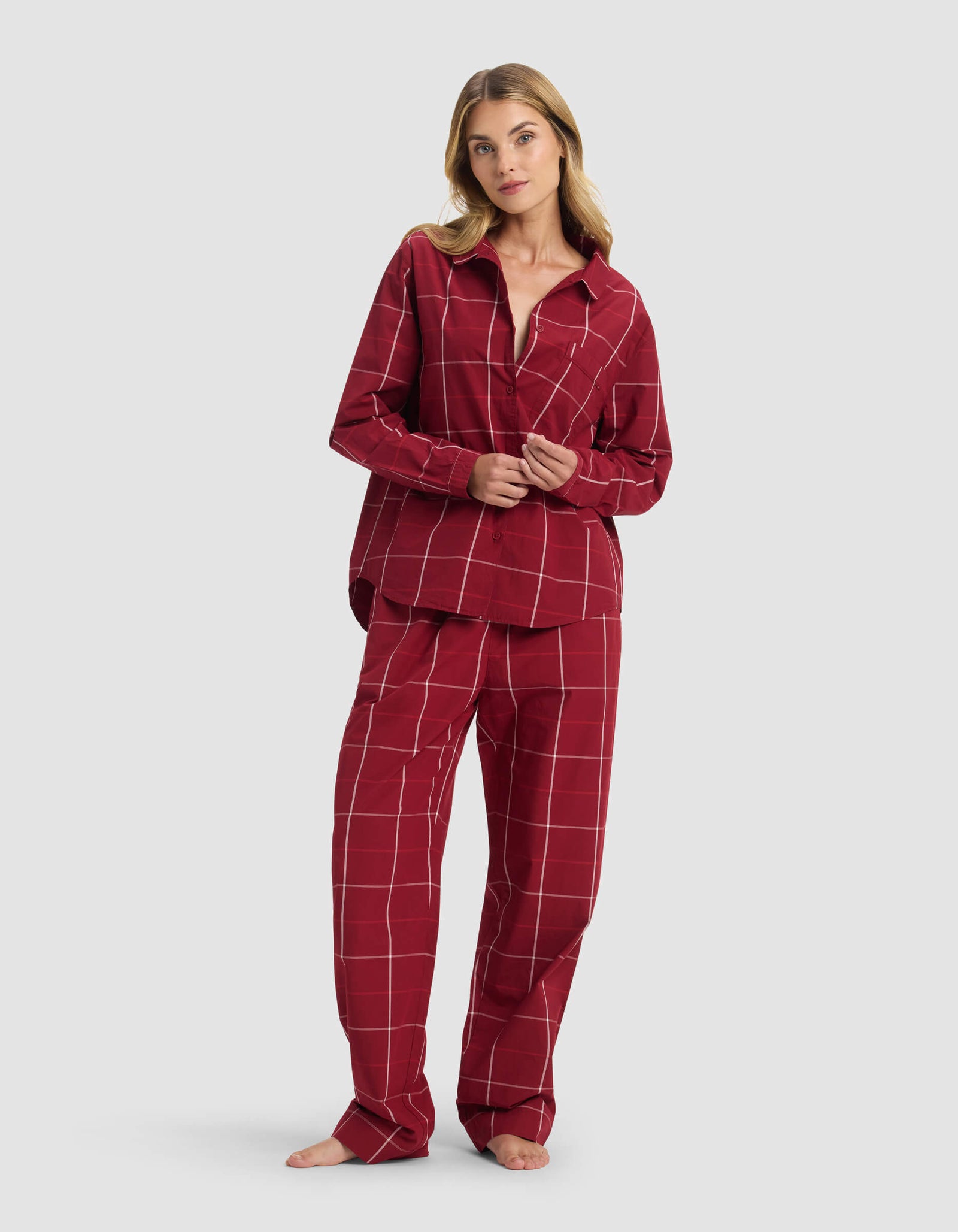 A woman stands barefoot against a light gray background, wearing the Cozy Earth Women's Sutton Long Sleeve Pajama Set—a matching red set with a white checkered pattern, featuring a button-up shirt and loose pants.