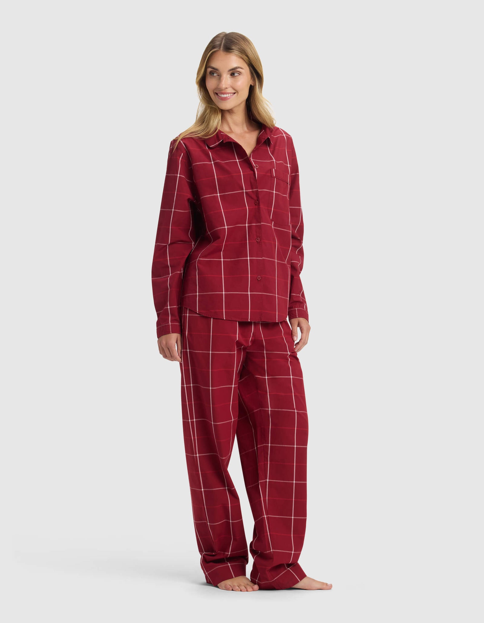A woman stands barefoot, smiling and looking to the side in the Cozy Earth Women’s Sutton Long Sleeve Pajama Set—matching red plaid pajamas with white lines, featuring a button-up shirt and loose-fit pants.