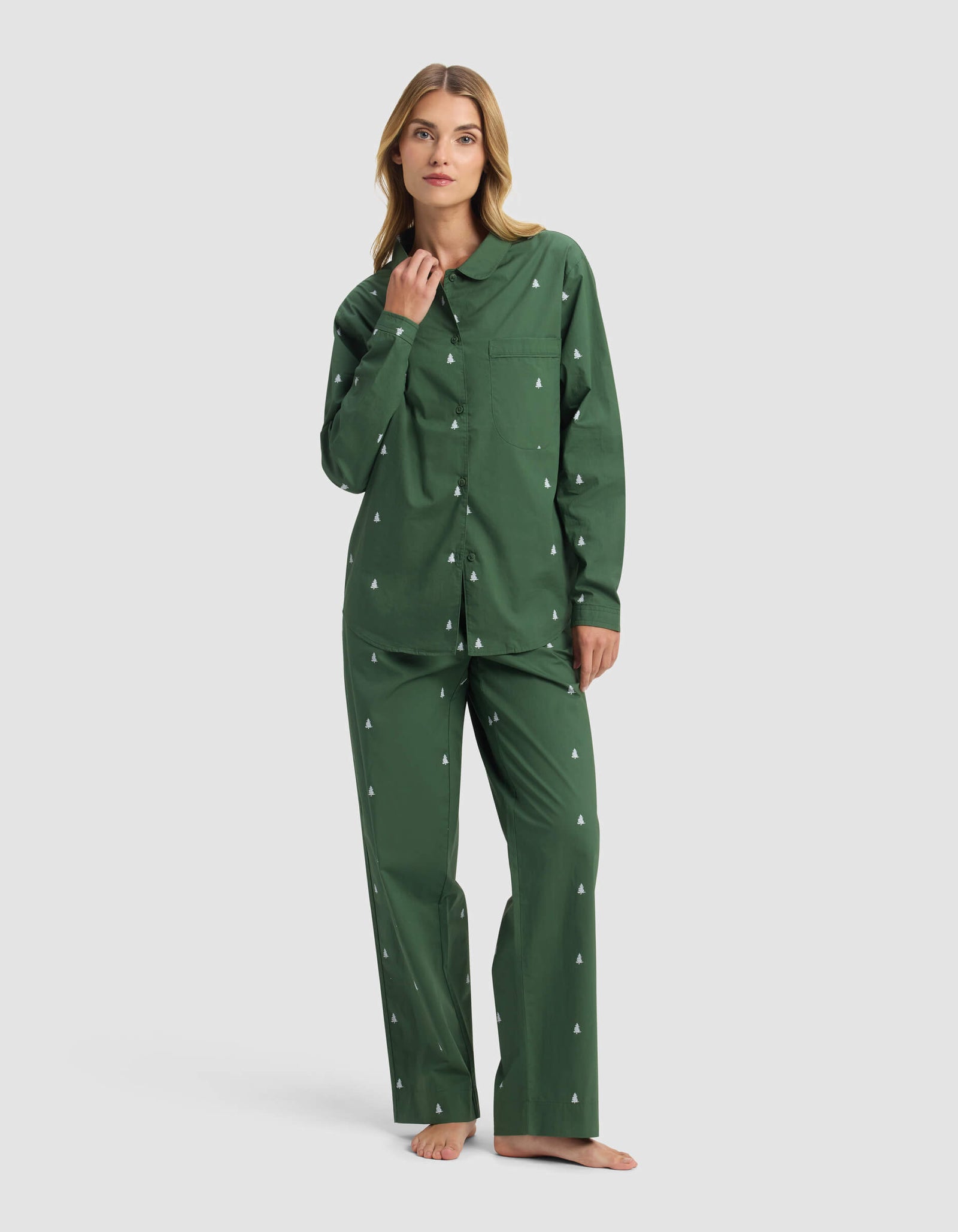A woman models the Cozy Earth Women's Sutton Long Sleeve Pajama Set, featuring a green button-up top and loose pants with white tree patterns. She stands barefoot, hand near her collar, and has a neutral expression.