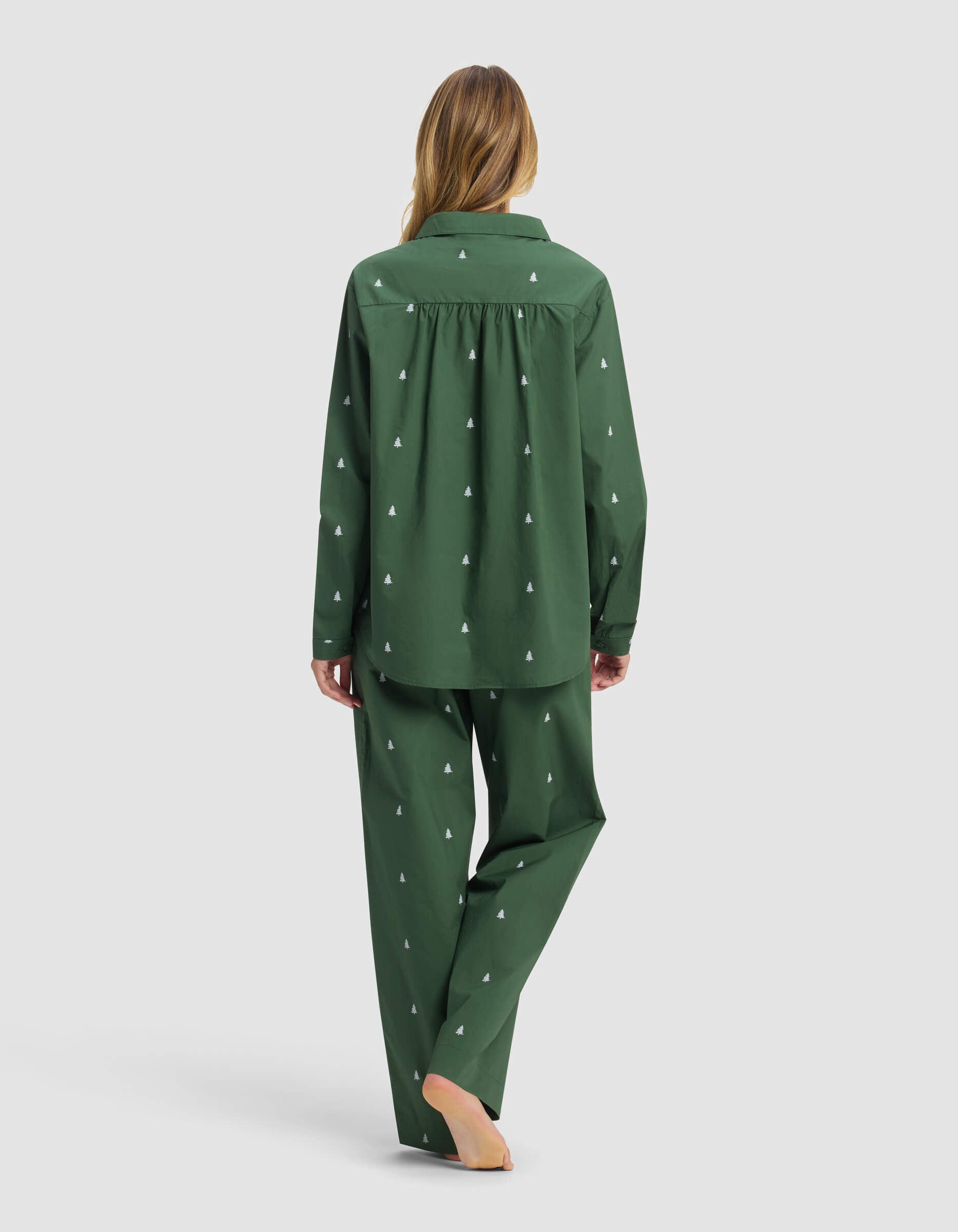 A woman with long hair stands barefoot, facing away, wearing the Cozy Earth Women's Sutton Long Sleeve Pajama Set in dark green with white tree patterns on both top and pants against a plain light background.|Color:Jade Tree